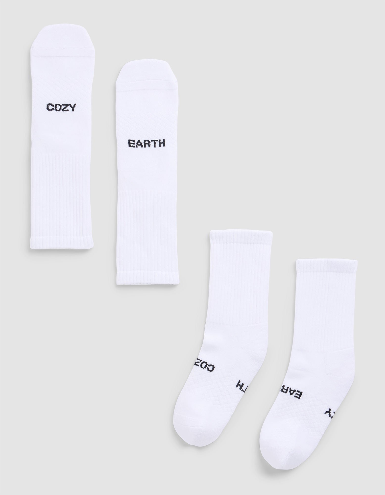 Four Essential Calf Socks by Cozy Earth are displayed on a light gray surface. Each white ribbed sock features black text: two say "COZY," and two say "EARTH." The socks are arranged flat and side by side. |Color:White
