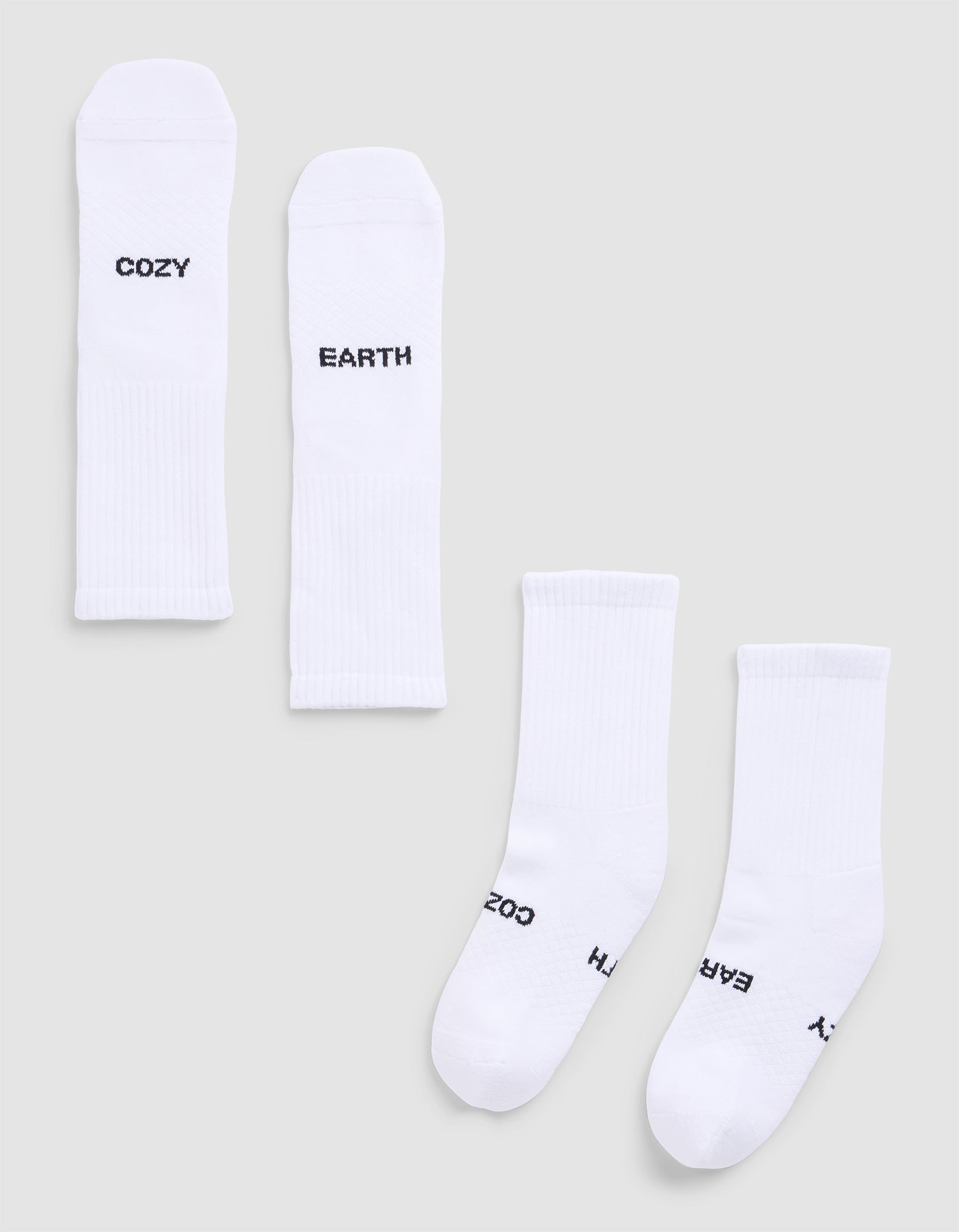 Four Essential Calf Socks by Cozy Earth are displayed on a light gray surface. Each white ribbed sock features black text: two say "COZY," and two say "EARTH." The socks are arranged flat and side by side. 