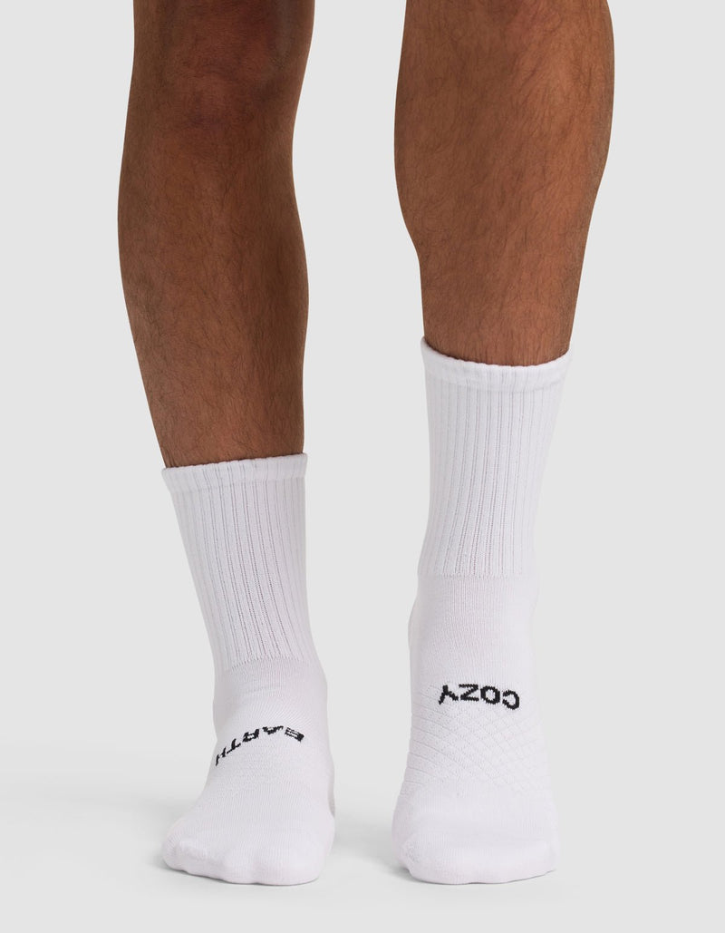 A person stands on a white surface wearing Essential Calf Socks by Cozy Earth. The white ribbed socks feature "COZY" and "EARTH" in black on the soles, visible from the back view.