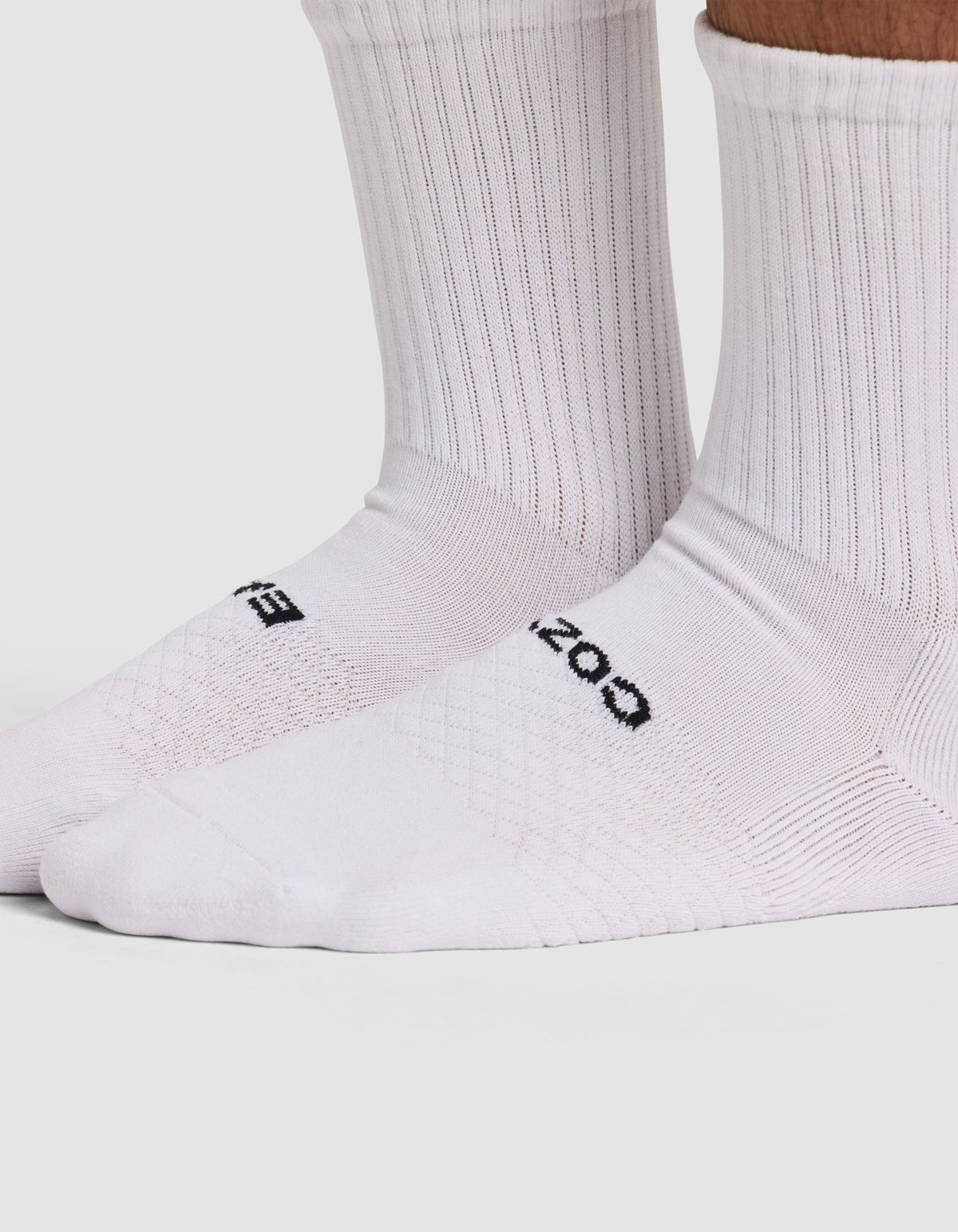 Close-up of a person wearing Cozy Earth Essential Calf Socks. These white ribbed socks feature black text on the top, cover the ankles and lower legs, and are set against a light, plain background.|Color:White