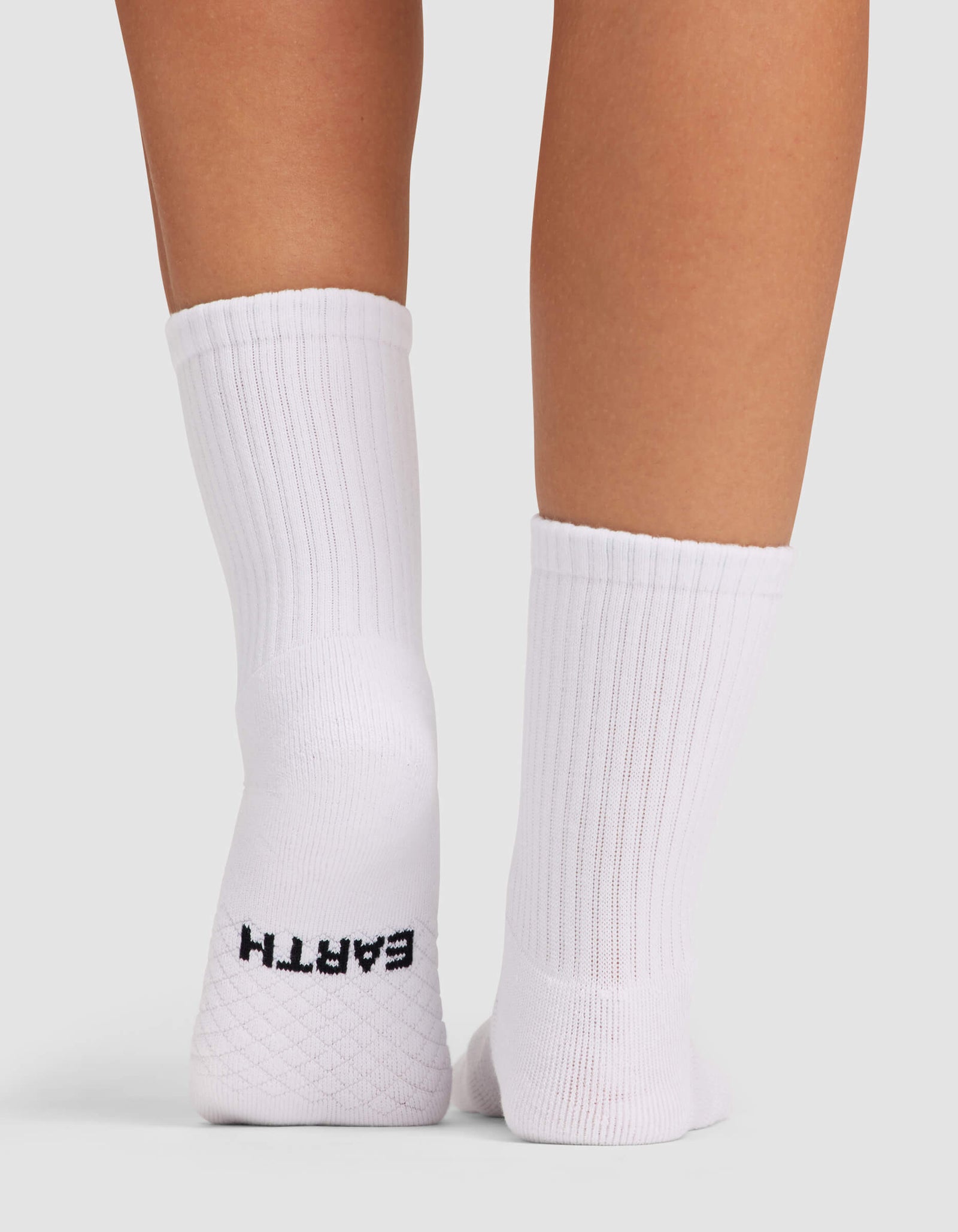 A close-up shows the back of two legs wearing Cozy Earth Women's Essential Calf Socks in white ribbed fabric. The left sock features "EARTH" in black on the heel, set against a plain light gray background.