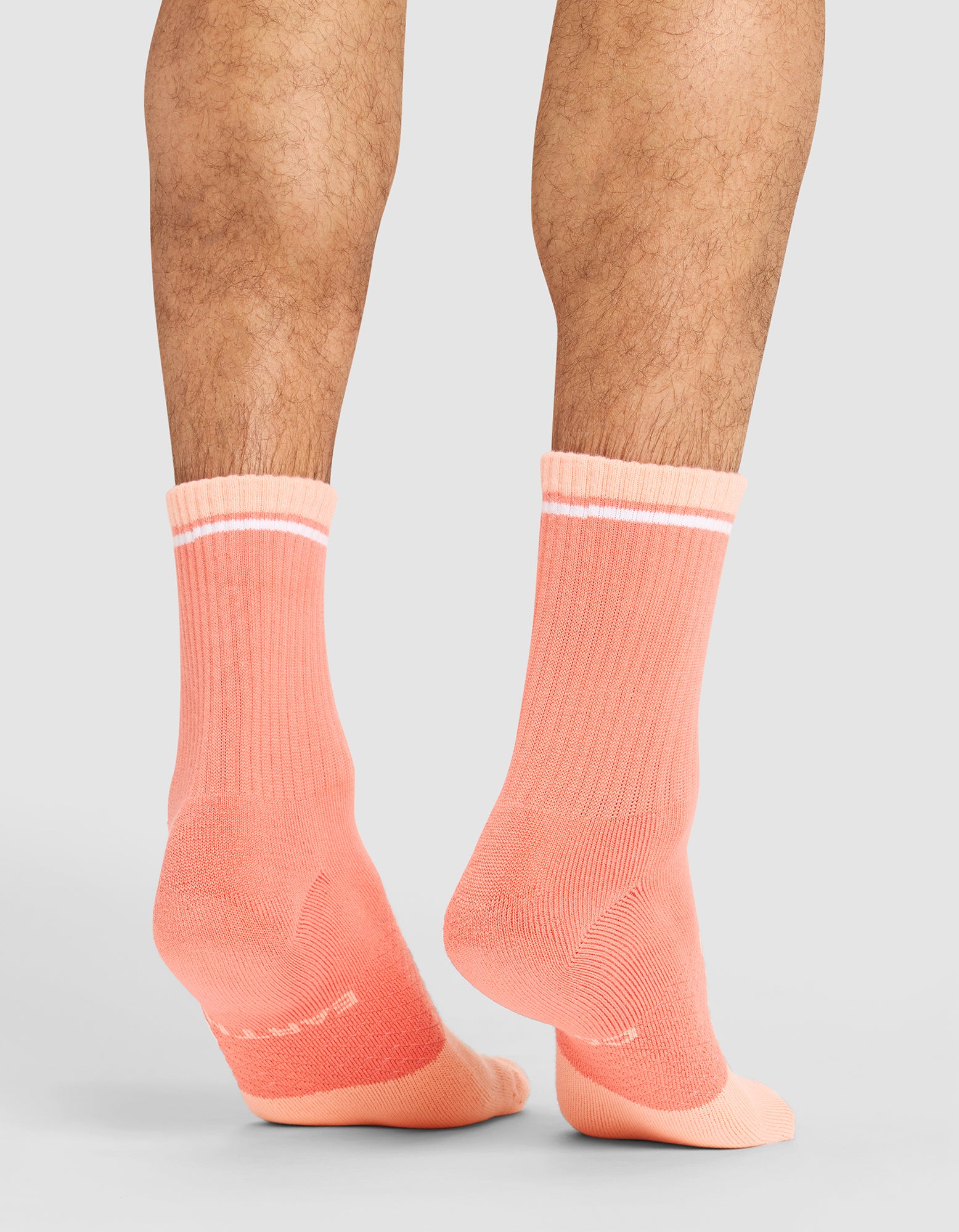 A person models Cozy Earth's Essential Calf Sock in coral with white top stripes, standing on a neutral background. The image highlights their lower legs and feet, showcasing the back and side views of the socks. 