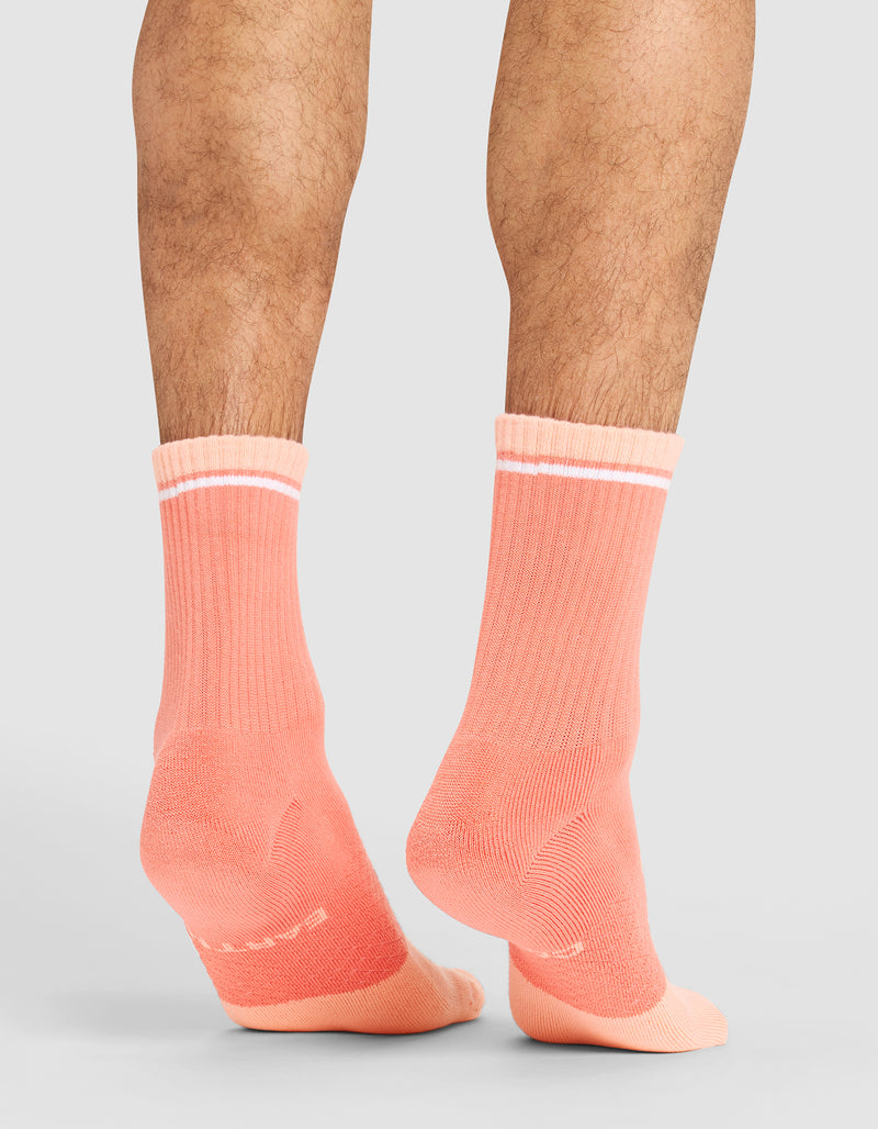 A person models Cozy Earth's Essential Calf Sock in coral with white top stripes, standing on a neutral background. The image highlights their lower legs and feet, showcasing the back and side views of the socks.