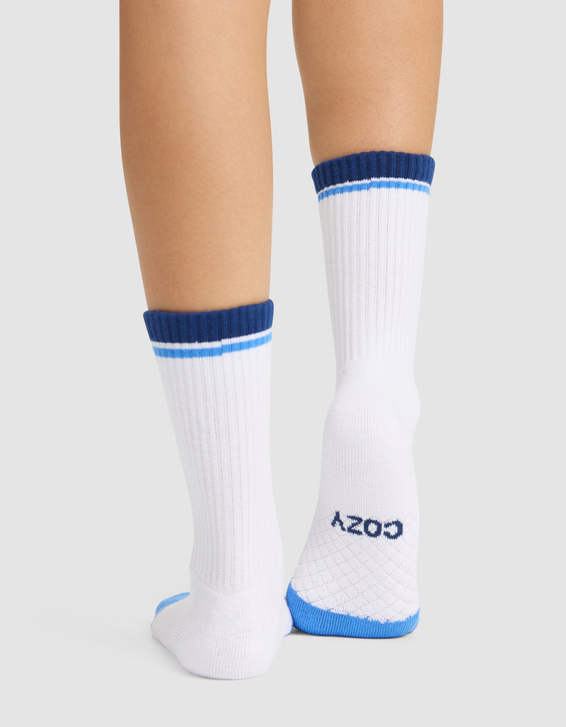 A person wears Cozy Earth Essential Calf Socks—white with blue stripes near the top, ribbed texture, and "COZY" on the bottom—standing on a light background.