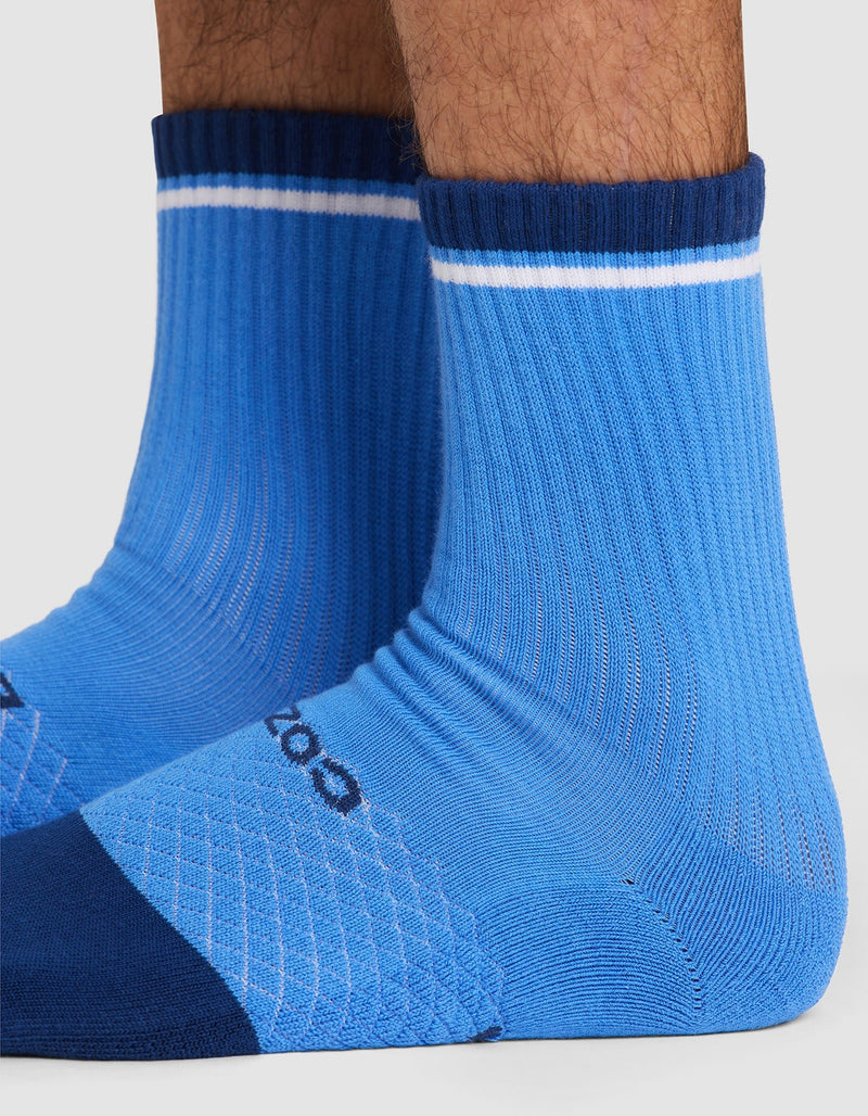 Close-up of a person's lower legs wearing Cozy Earth Essential Calf Socks—bright blue ribbed crew socks with dark blue toes and heels, a white stripe near the top, and a small side logo.