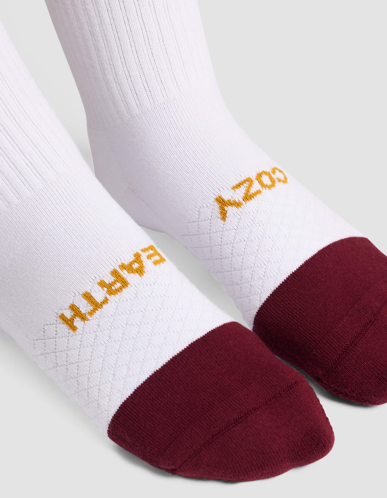 The Women's Essential Calf Sock by Cozy Earth features white fabric, maroon toes, "COZY EARTH" in gold on top, a ribbed upper section, and a textured midfoot pattern.