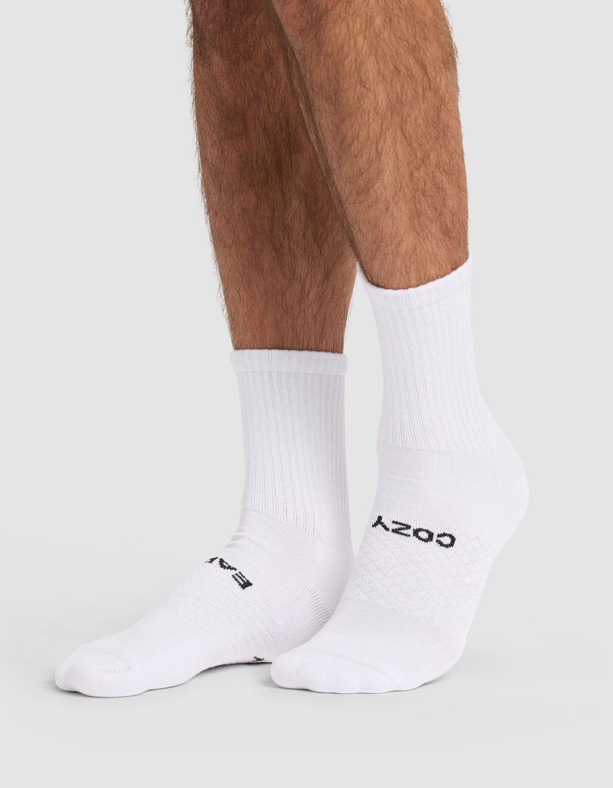 Someone wears Cozy Earth Essential Calf Sock 4-Pack—white crew socks with black text, covering ankles and lower legs—while standing on a light gray surface.
