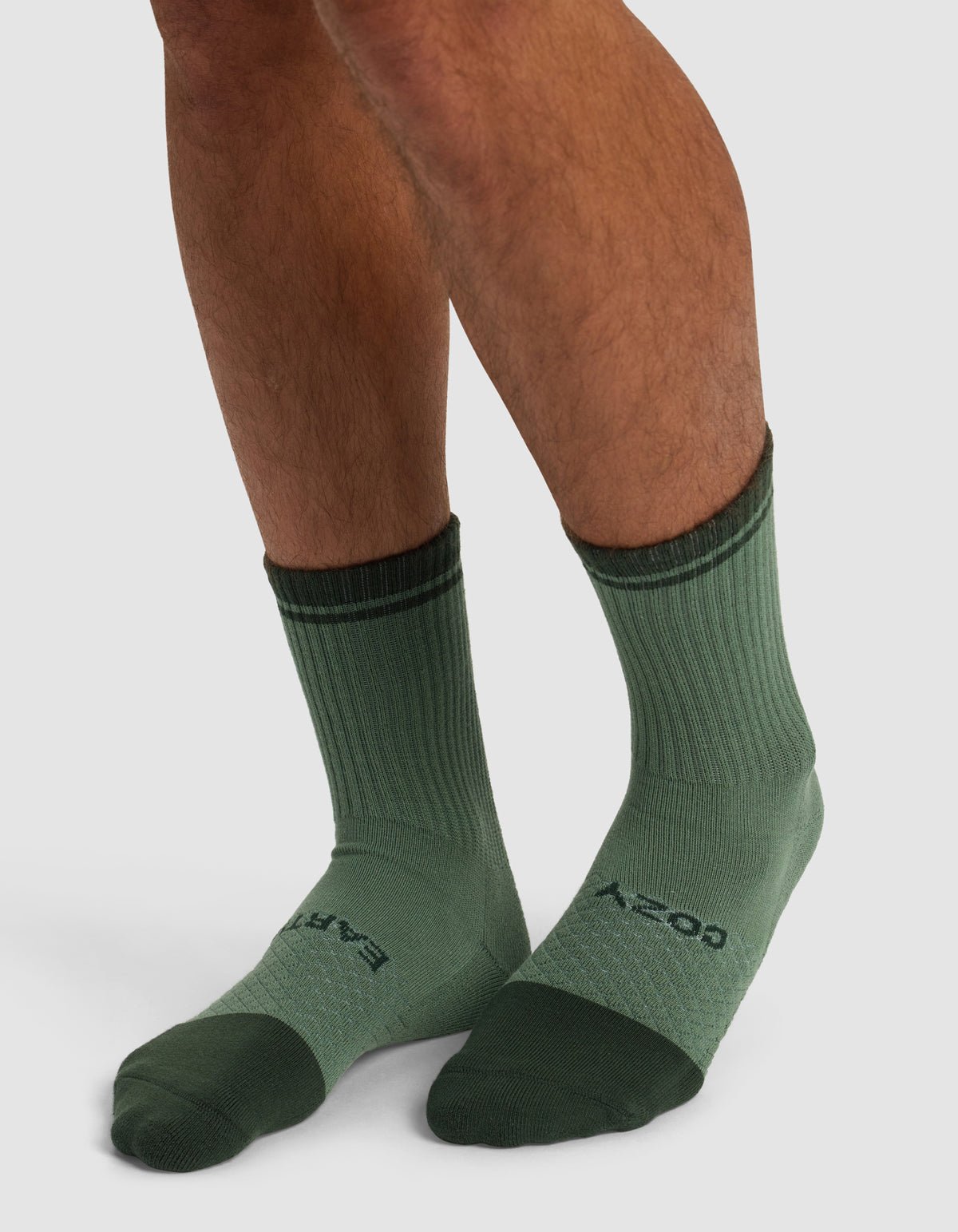 Close-up of a person's lower legs wearing Cozy Earth Essential Calf Sock - Last Chance in green, featuring darker green heels, toes, and cuffs. The socks have text on the top of the foot and are set against a plain light background.