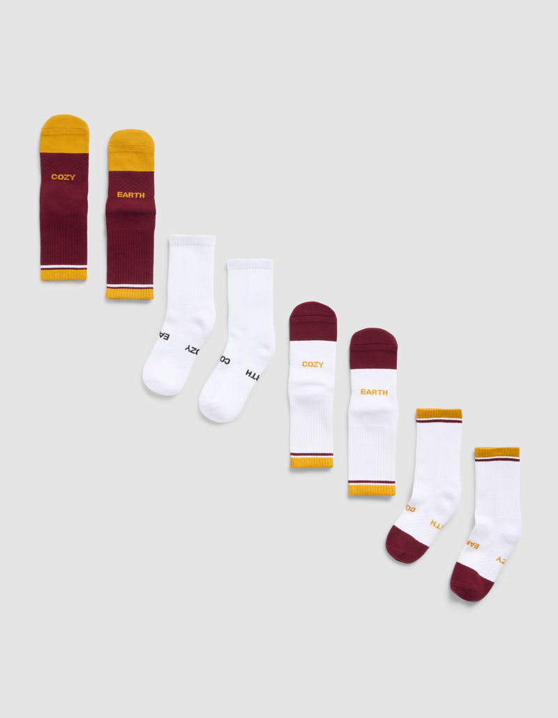 The Essential Calf Sock 4-Pack by Cozy Earth features four pairs of socks in maroon and yellow, plain white, maroon with white and yellow accents, and white with colorful details—some decorated with words like “COZY” and “EARTH.”.