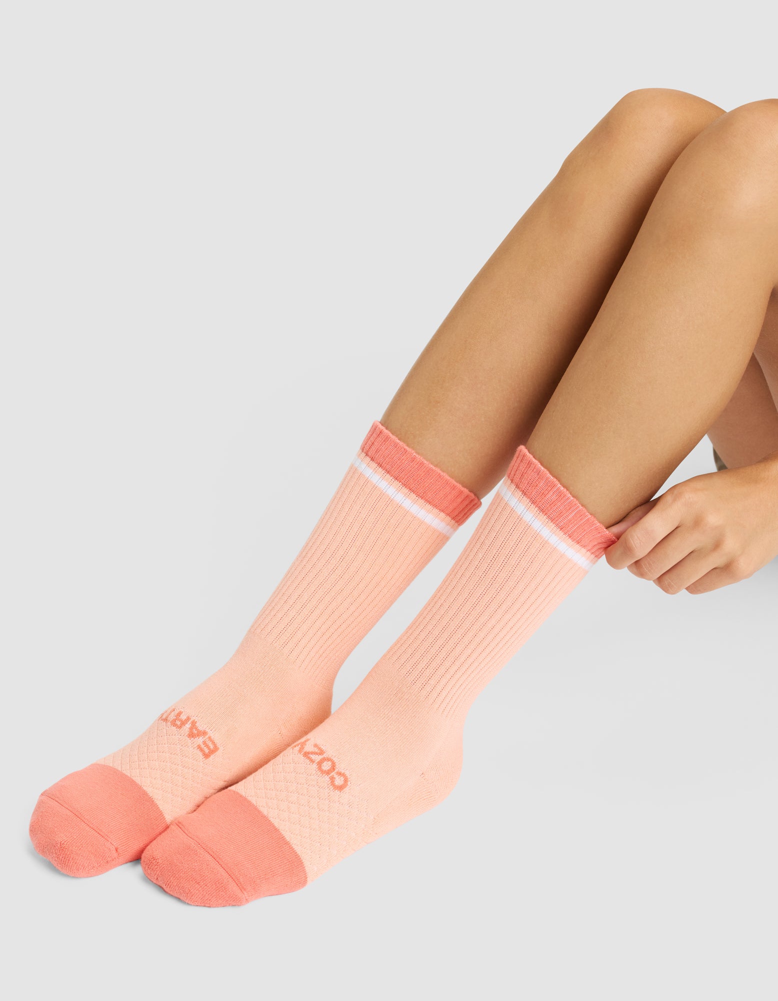 A person sits on the floor wearing Essential Calf Socks by Cozy Earth—peach crew socks with coral toes, heels, and cuffs, a white stripe near the top, and text on the toes. Only their legs and hands are visible. 