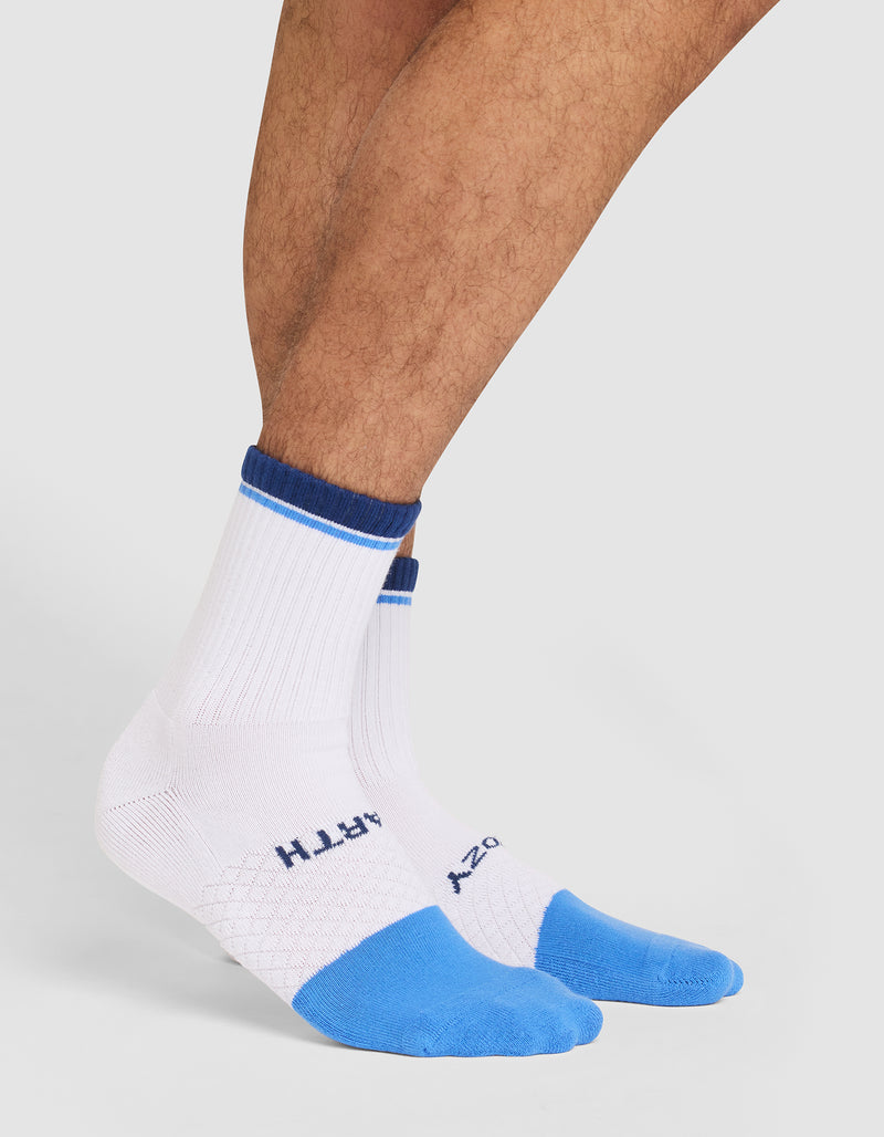 A person wearing Cozy Earth Essential Calf Socks—white crew socks with blue toes, heels, a blue top stripe, and text near the toes. Only the lower legs and feet are visible against a plain background.