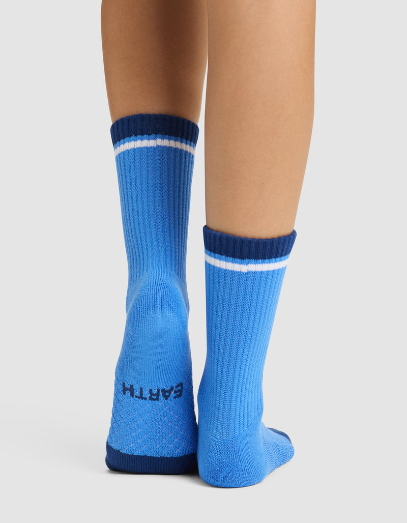 A person wears Cozy Earth's Essential Calf Sock in bright blue with dark blue cuffs and a white stripe. "EARTH" is printed on the bottom of the left sock. The back view highlights the heel and cuffs.