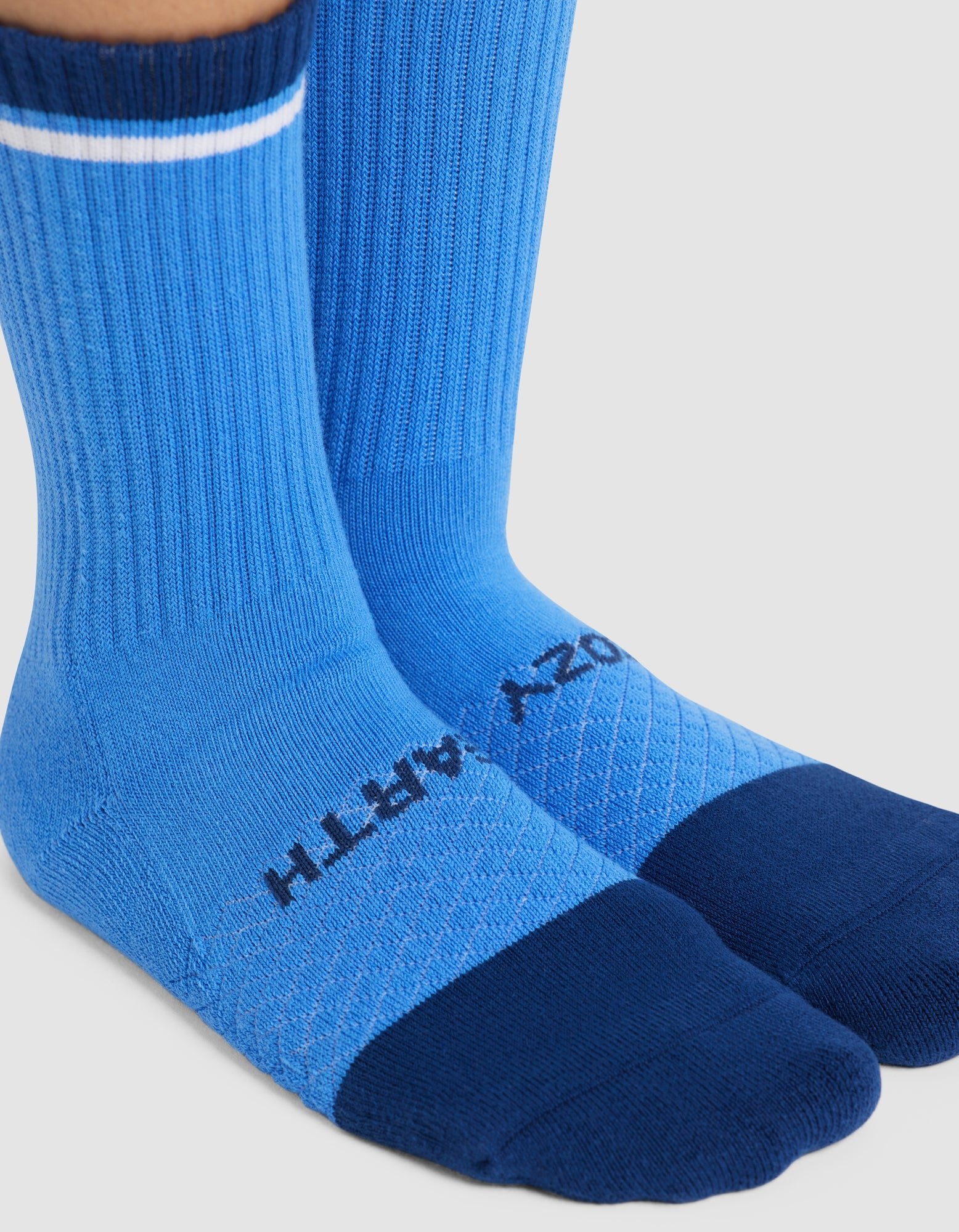 A pair of feet wearing Cozy Earth Essential Calf Socks in blue, featuring a darker blue toe and heel, a white stripe near the top, and "COZY EARTH" printed in black on the top of the foot. 