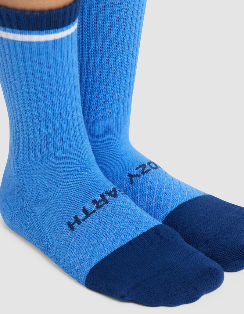 A pair of feet wearing Cozy Earth Essential Calf Socks in blue, featuring a darker blue toe and heel, a white stripe near the top, and "COZY EARTH" printed in black on the top of the foot.