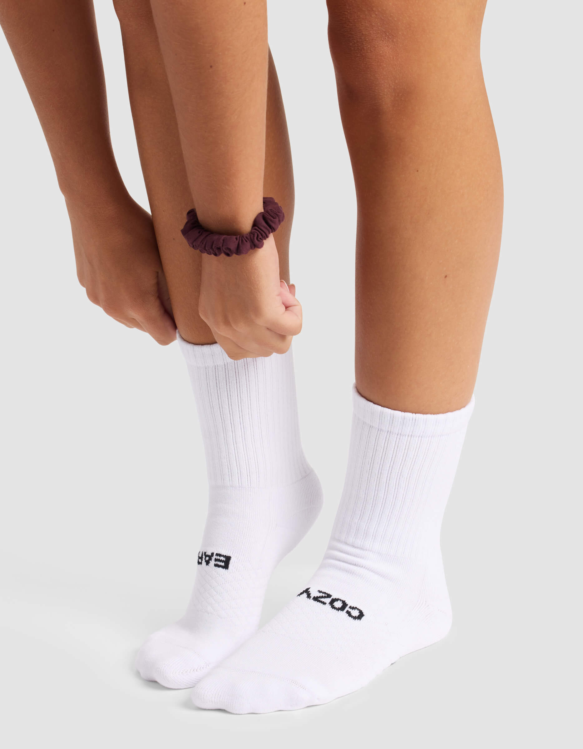 A person wearing Cozy Earth's Women's Essential Calf Sock in white, featuring "COZY" and "EASY" text, adjusts one sock. They have a dark scrunchie on their wrist and stand on a light surface.|Color:White
