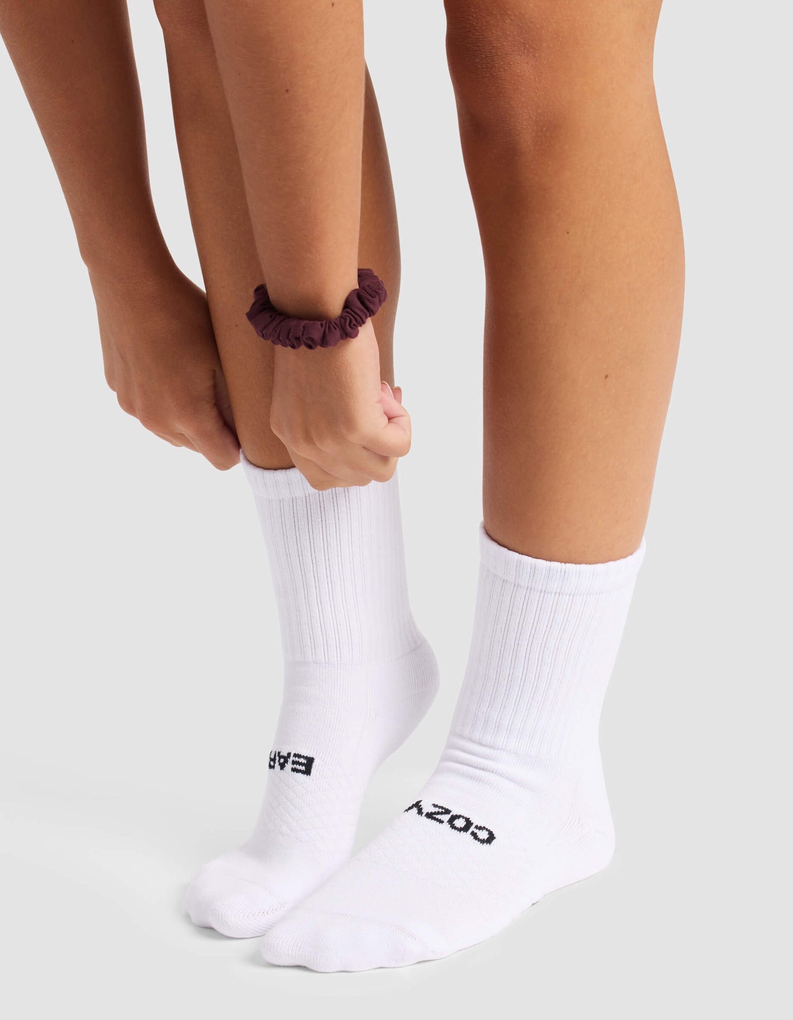 A person wearing Cozy Earth's Women's Essential Calf Sock in white, featuring "COZY" and "EASY" text, adjusts one sock. They have a dark scrunchie on their wrist and stand on a light surface.