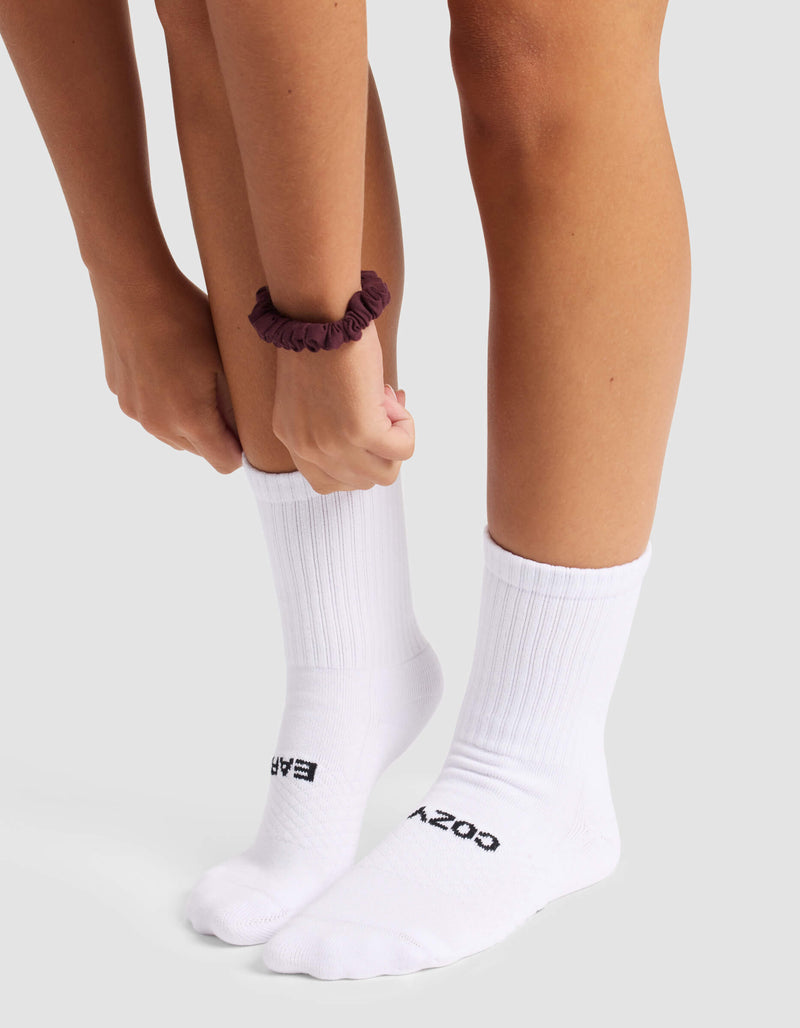 A person wearing Cozy Earth's Women's Essential Calf Sock in white, featuring "COZY" and "EASY" text, adjusts one sock. They have a dark scrunchie on their wrist and stand on a light surface.