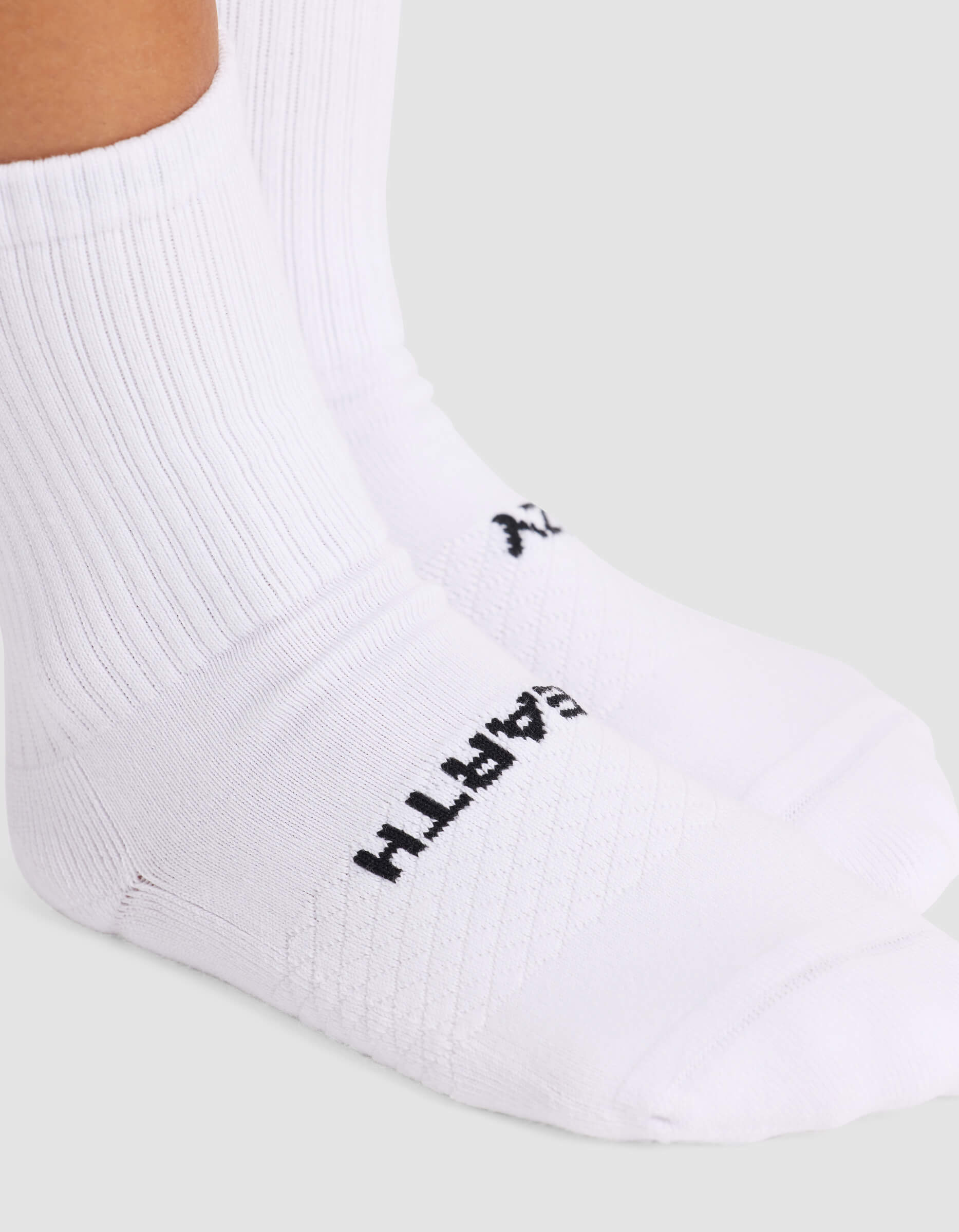 A close-up shows a person wearing Cozy Earth Women’s Essential Calf Socks in white with ribbing and a black logo near the toes. The background is plain and light-colored.