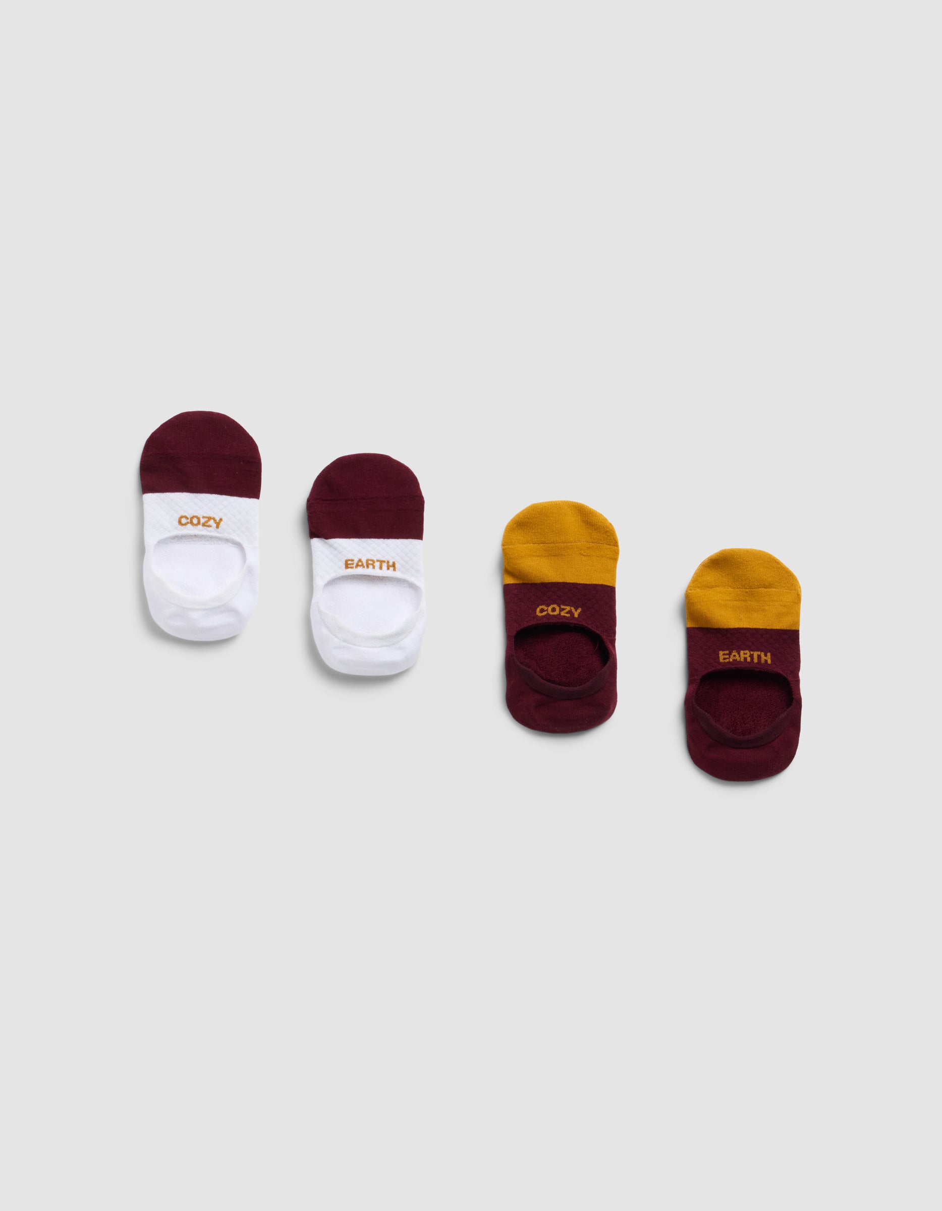 Four pairs of Essential No-Show Socks by Cozy Earth are arranged in a row on a light gray background: two pairs in white and maroon, and two in maroon and mustard yellow, each with "COZY" and "EARTH" text.|Color:Burgundy