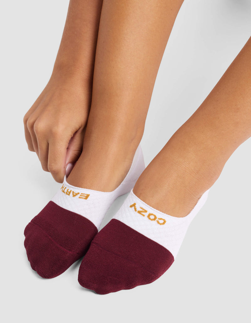 A person adjusts Cozy Earth's Women's Essential No-Show Socks in white and burgundy on their feet; “COZY” and “EARTH” appear in gold lettering near the toes.