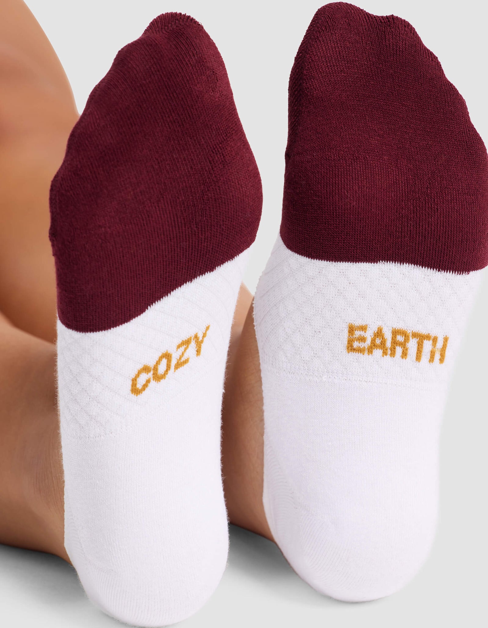 A close-up of two feet wearing Cozy Earth Women's Essential No-Show Socks in white with maroon toes. The soles display "COZY" on the left and "EARTH" on the right in yellow letters.