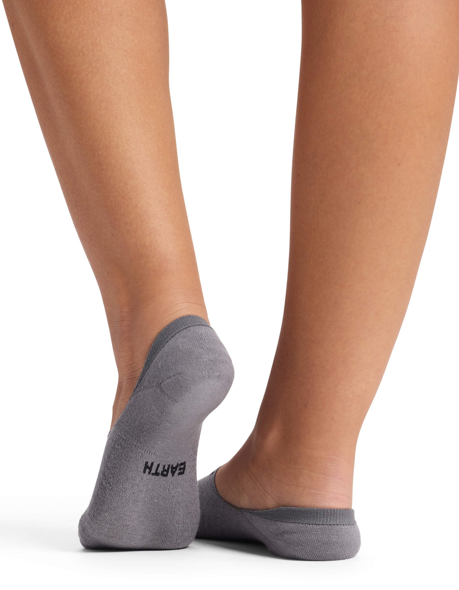 A woman stands with one foot crossed behind the other, showing only her lower legs and feet in Cozy Earth Women's Essential No-Show Socks, low-cut and gray, with "EARTH" printed on the sole against a white background.