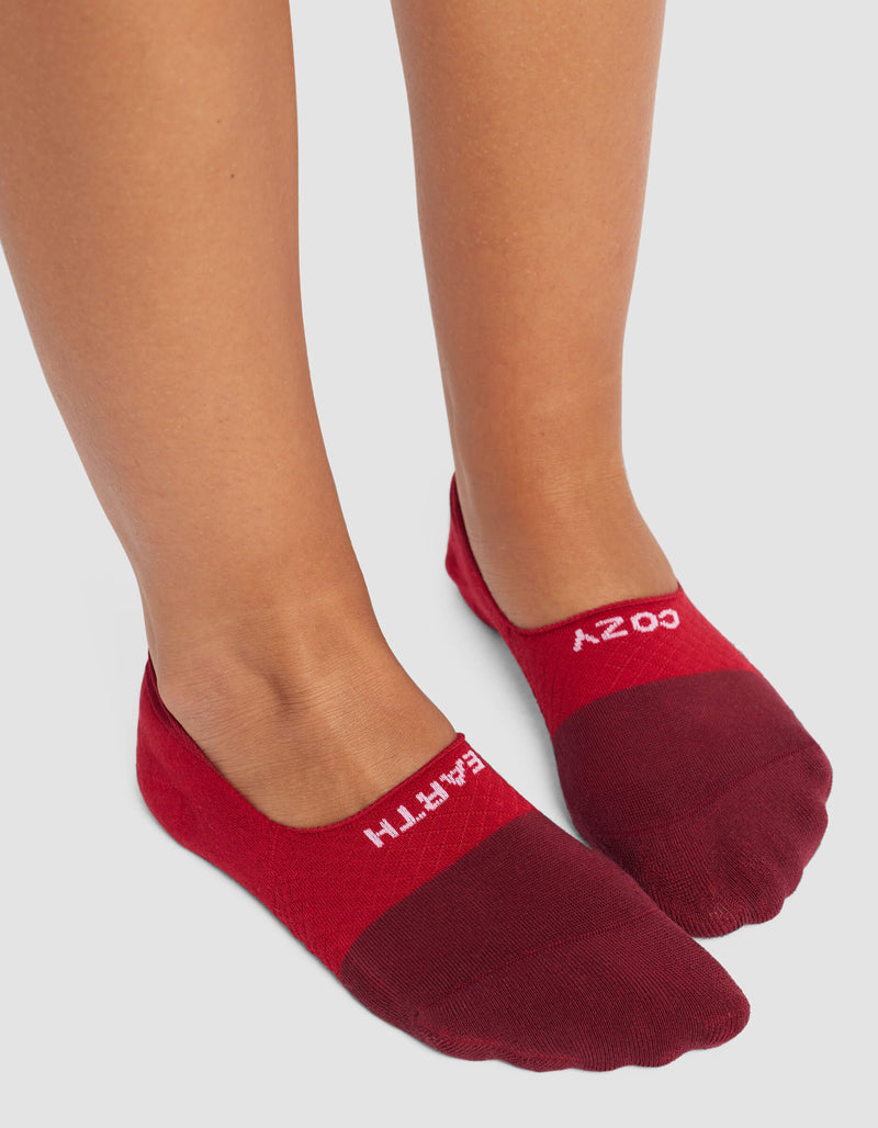 A person stands on a light gray surface wearing maroon and red Women's Essential No-Show Socks by Cozy Earth, featuring "COZY EARTH" in white text.