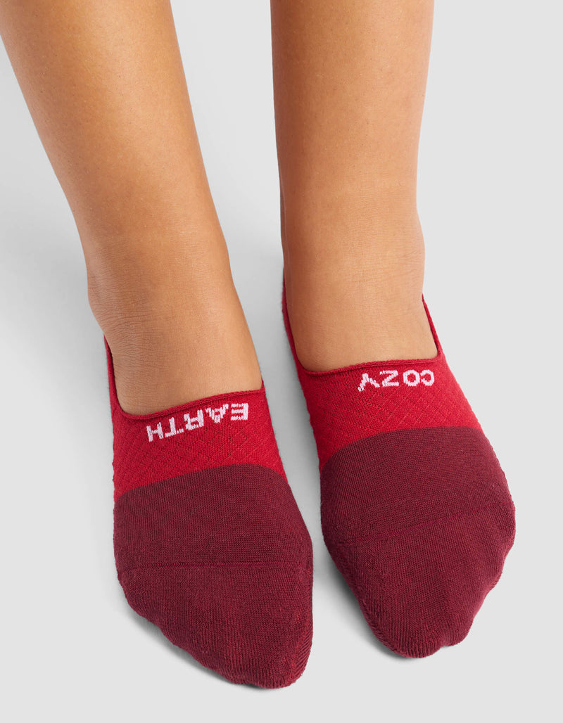A person stands on a light gray surface wearing Cozy Earth's Women's Essential No-Show Socks in maroon, featuring "EARTH" on the left sock and "COZY" on the right.