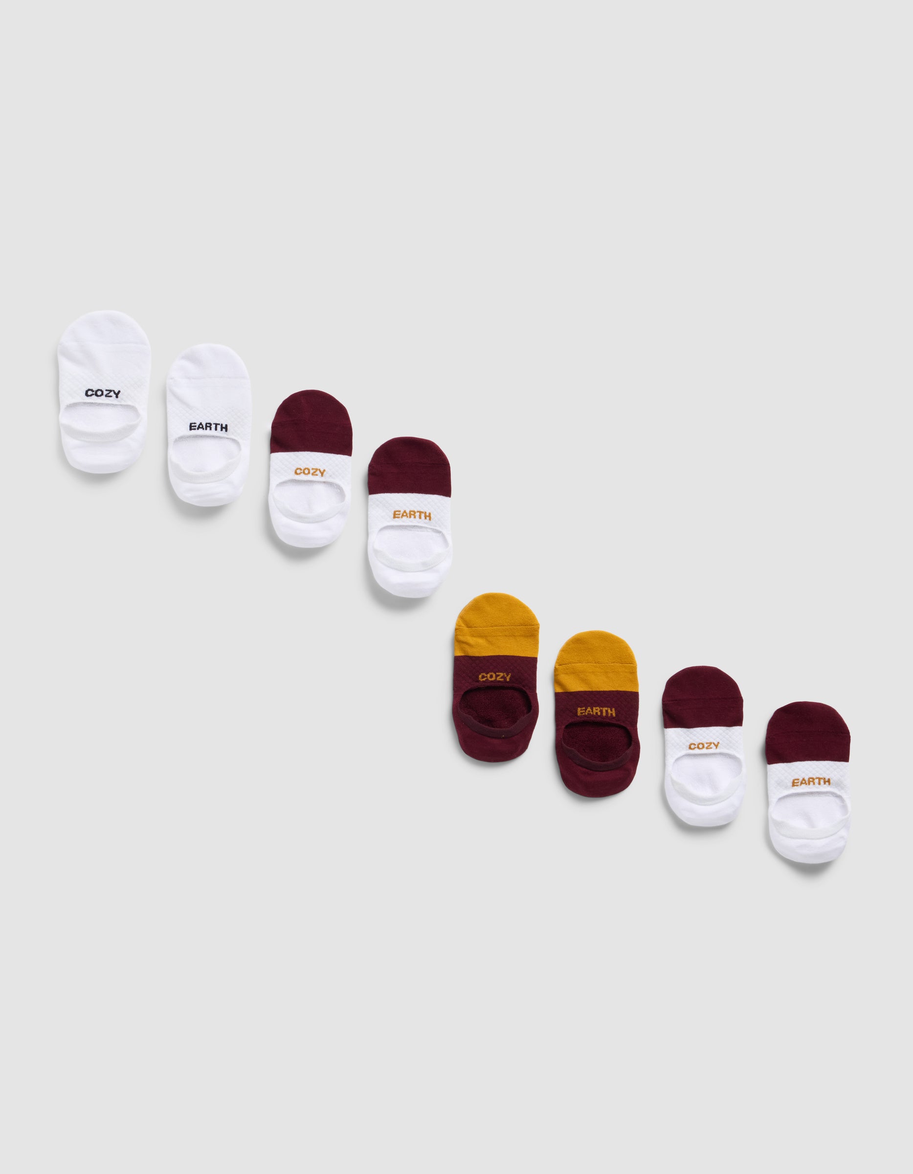 Eight pairs of Cozy Earth Essential No-Show Socks in white, burgundy, and yellow are arranged diagonally on a light gray background. Some pairs feature "COZY" or "EARTH" printed on the cuffs.|Color:Burgundy