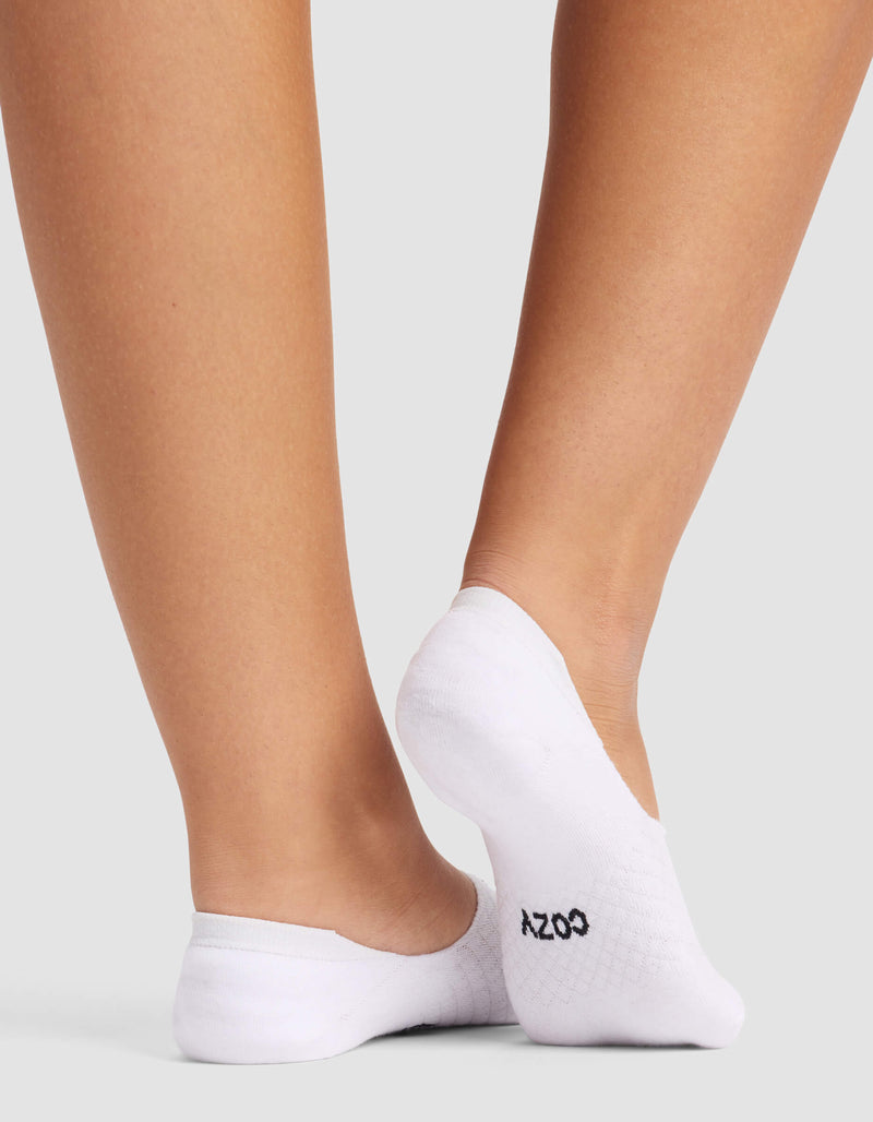 A person stands on a light gray surface wearing Cozy Earth’s Essential No-Show Sock, with "COZY" printed in black on the sole of the right sock, revealing the back of their lower legs and feet.