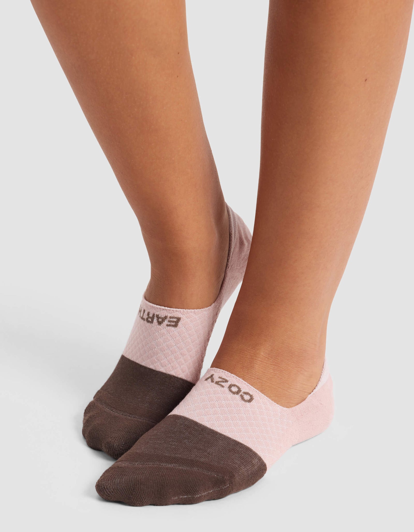 A person stands on a light gray surface wearing Cozy Earth Women's Essential No-Show Socks—low-cut, two-tone with brown toes, pale pink heels, and "COZY" on one sock, "EARTH" on the other.