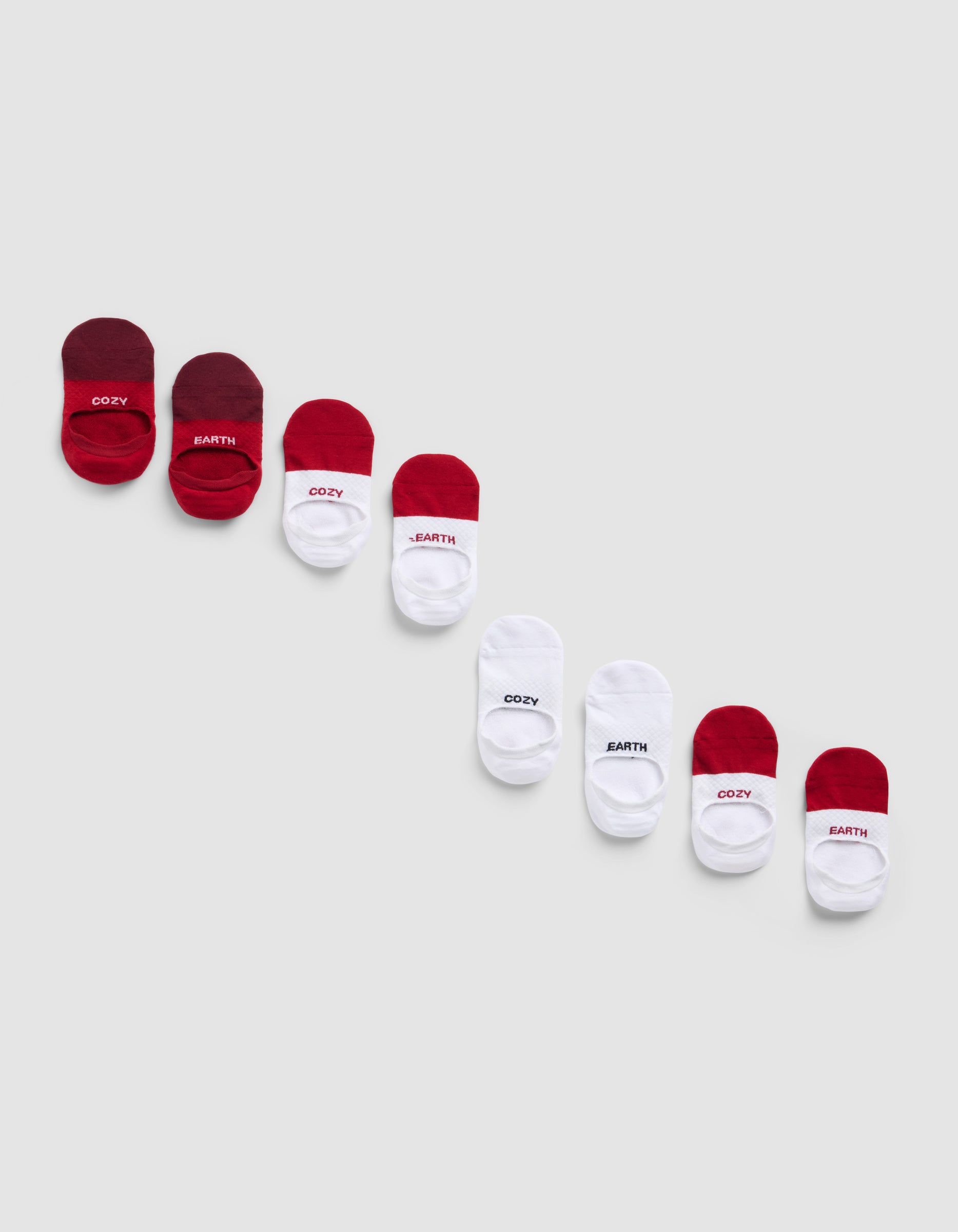 Four pairs of Essential No-Show Socks by Cozy Earth in red, white, and maroon are arranged diagonally on a light gray background, with some socks featuring "COZY" or "EARTH" printed on them.|Color:Crimson