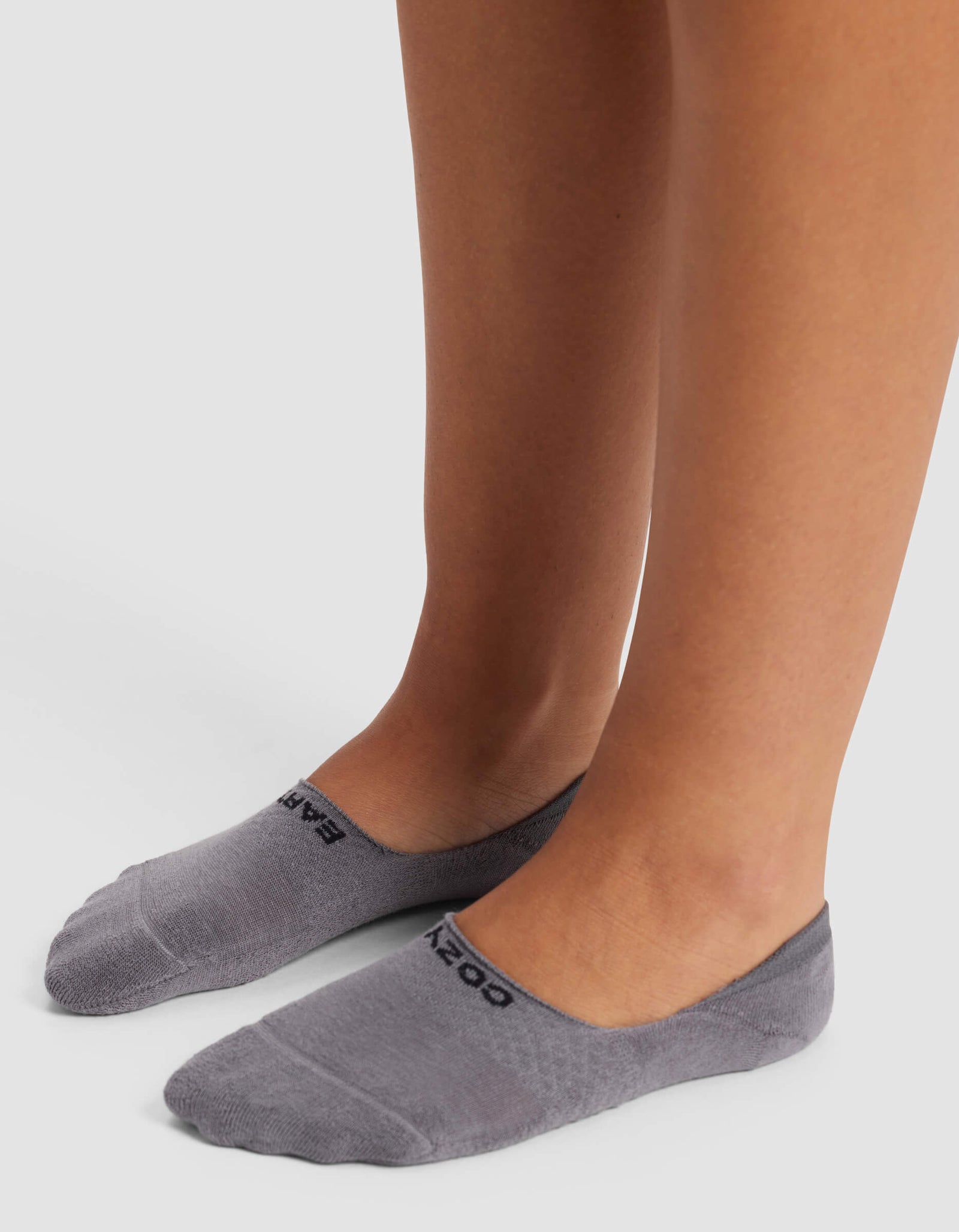 A person wears Cozy Earth Women's Essential No-Show Socks in gray, standing on a white surface. "COZY" appears near the toes on each sock.