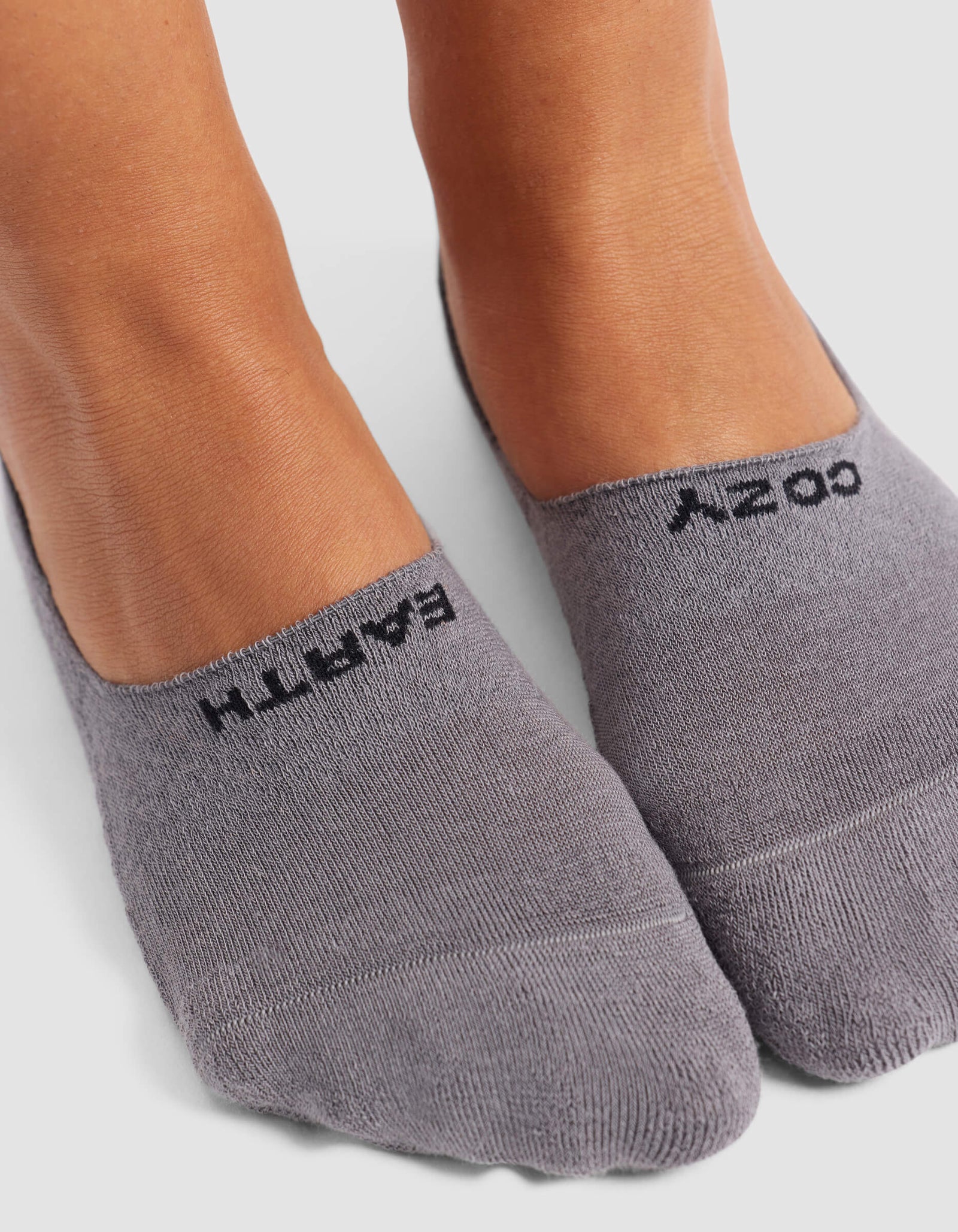 Close-up of feet wearing Cozy Earth Women's Essential No-Show Socks in light gray with “EARTH” and “COZY” printed in black on top, set against a plain, neutral background.
