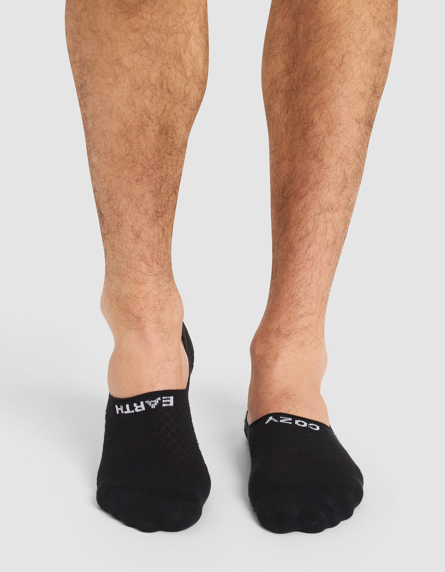 A person with bare legs wears Cozy Earth Essential No-Show Socks in black, featuring "EARTH" on the left and "COZY" on the right in white text. The background is plain and light-colored. |Color:Black