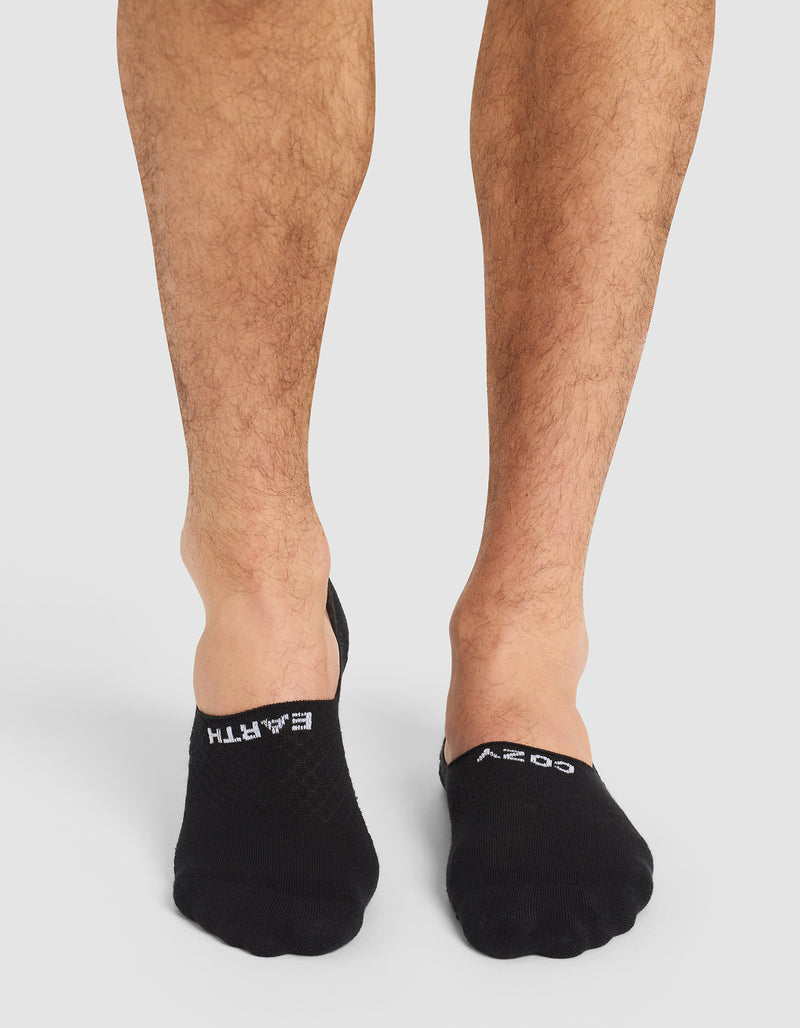 A person with bare legs wears Cozy Earth Essential No-Show Socks in black, featuring "EARTH" on the left and "COZY" on the right in white text. The background is plain and light-colored.