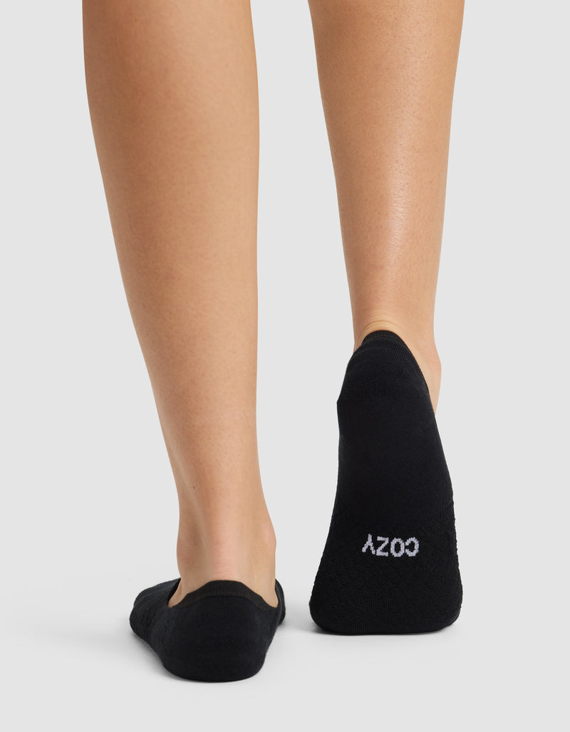 A person is seen from behind wearing Cozy Earth’s Essential No-Show Socks in black. "COZY" is printed in white on the sole of the left sock. The background is plain and light-colored.