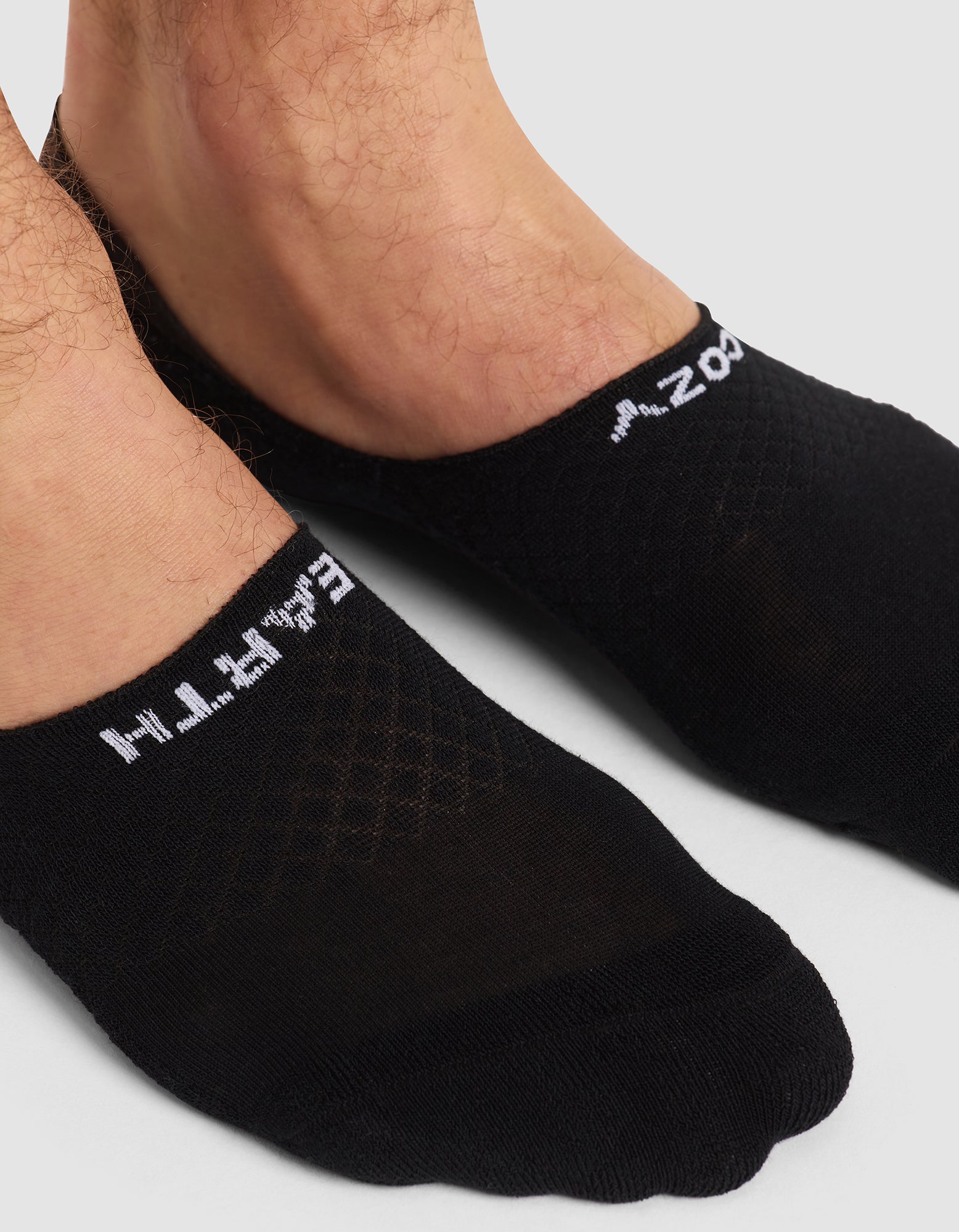 Close-up of feet wearing Cozy Earth's Essential No-Show Sock in black with white text near the ankle, standing on a light surface. 