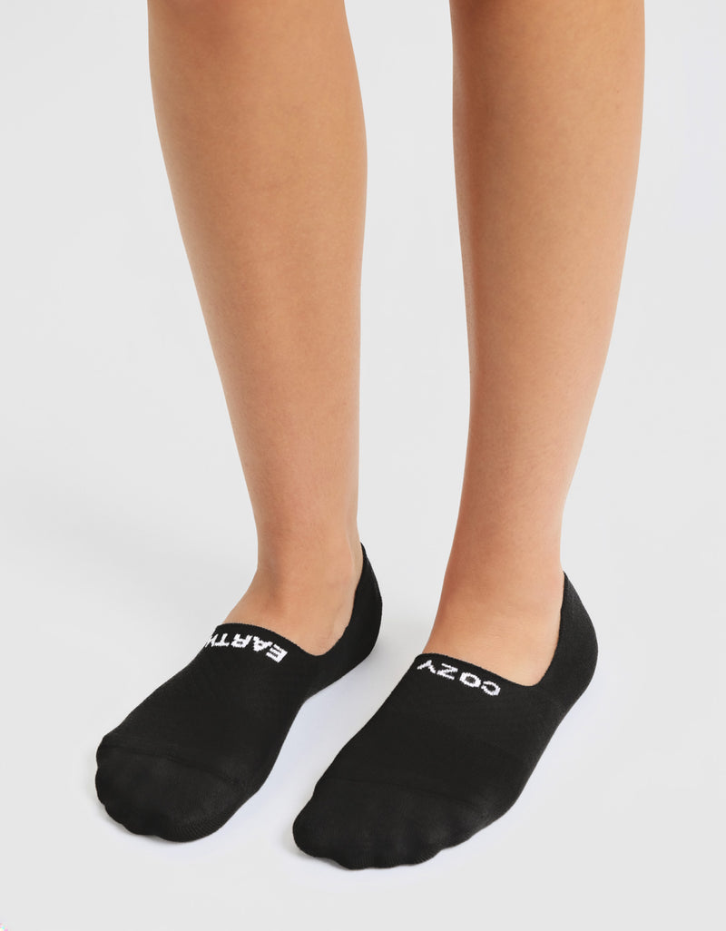 A person stands on a white surface wearing Cozy Earth Women's Essential No-Show Socks in black, with "EARTH" in white on the left sock and "COZY" in white on the right.