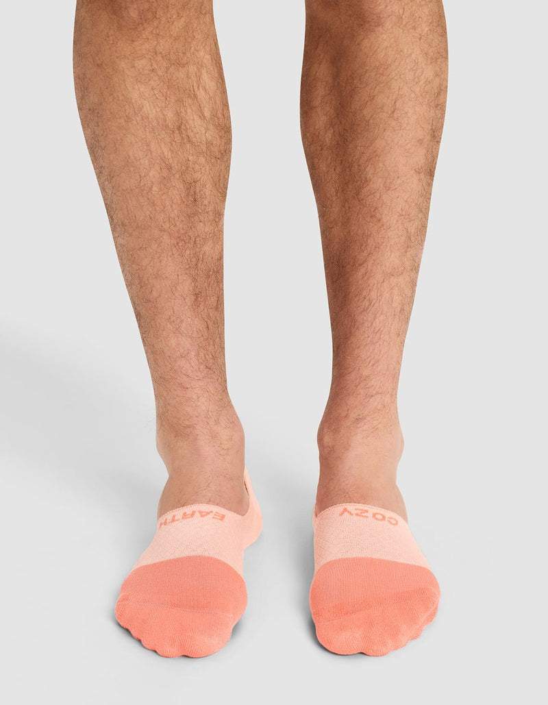 A person wearing Cozy Earth Essential No-Show Socks in light pink with peach toes, featuring text near the toes, stands on a light gray surface. Only their lower legs and feet are visible.