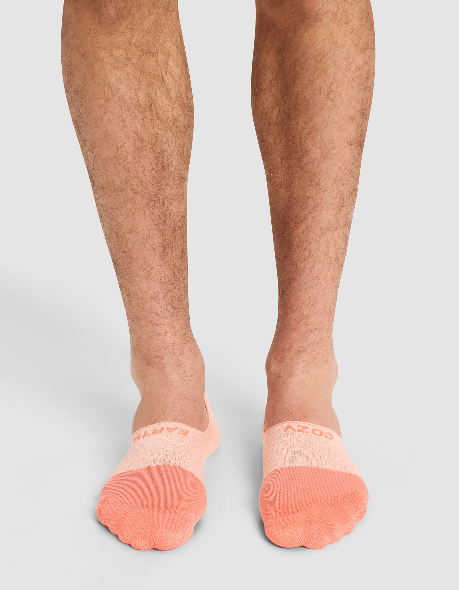A person with light to medium skin tone stands wearing Cozy Earth Essential No-Show Socks in peach, featuring "COZY EARTH" on top. The socks cover the feet and ankles against a plain light gray background. 