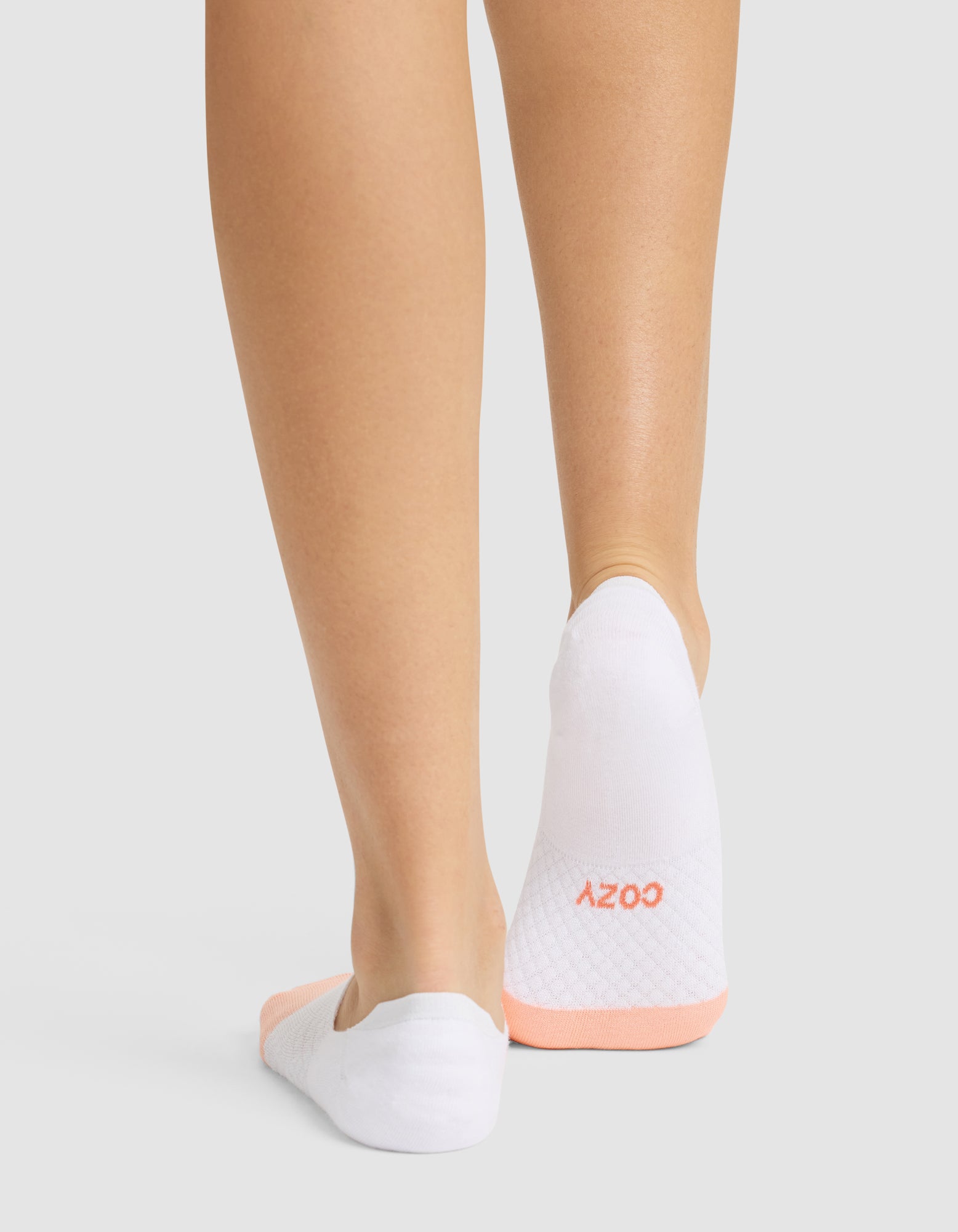 Back view of a person wearing Cozy Earth Essential No-Show Socks, featuring a peach-colored grip and "COZY" in peach on one sock, standing on a light gray surface. 