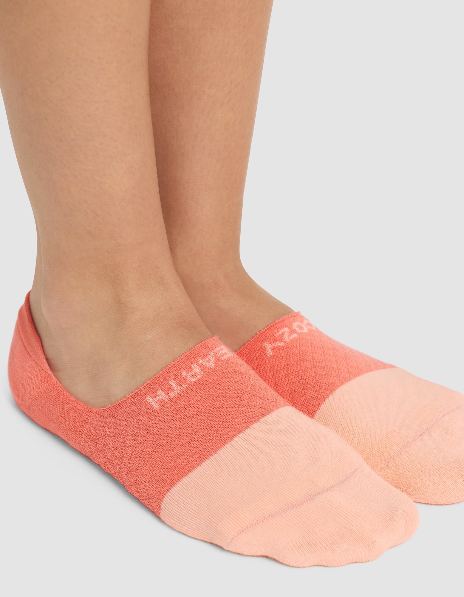 A close-up of two feet wearing Cozy Earth’s Essential No-Show Socks in peach and coral, featuring partially visible white "CRAZY EARTH" text near the ankle, standing on a light gray surface. |Color:Cantaloupe
