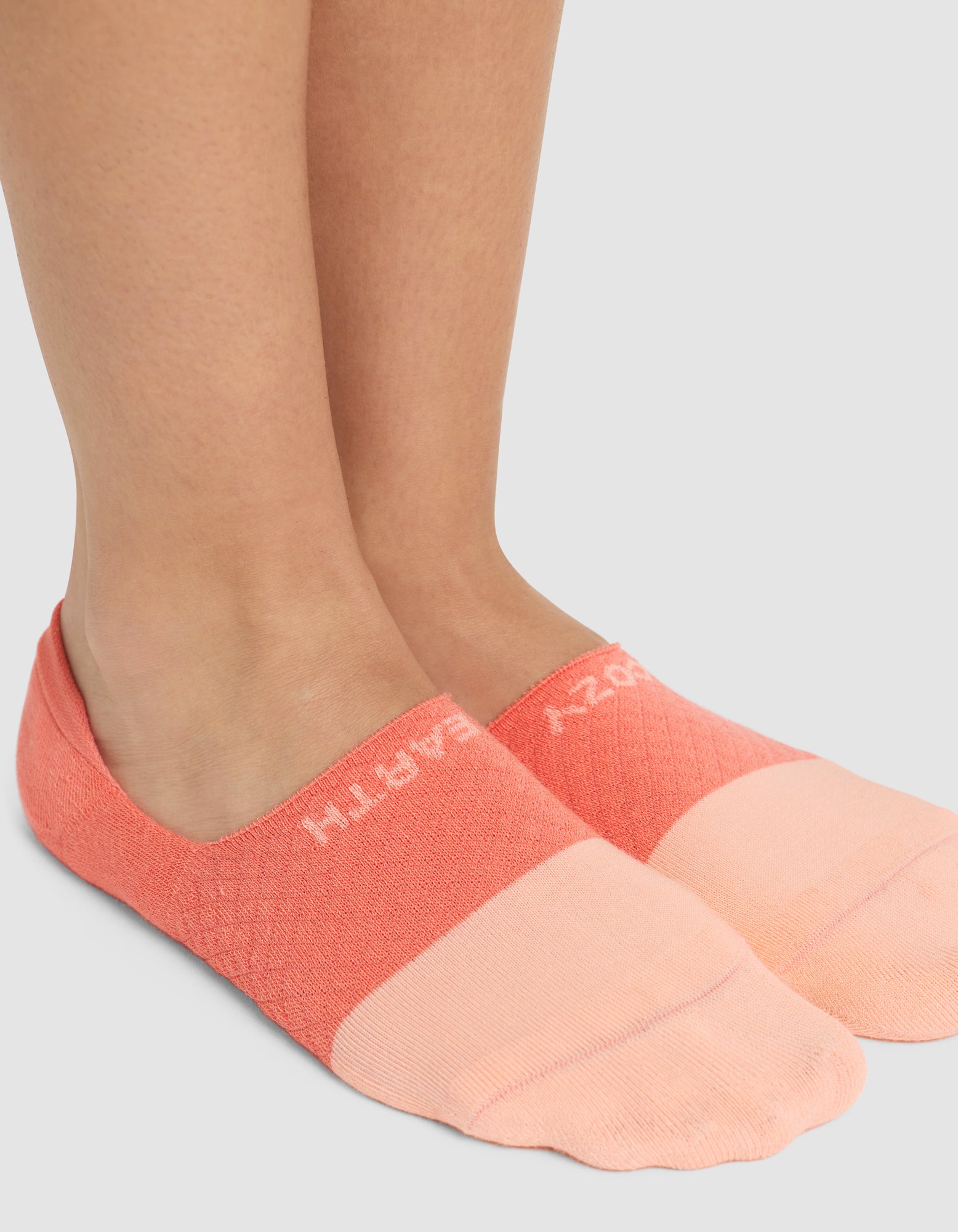 A close-up of two feet wearing Cozy Earth’s Essential No-Show Socks in peach and coral, featuring partially visible white "CRAZY EARTH" text near the ankle, standing on a light gray surface. |Color:Cantaloupe