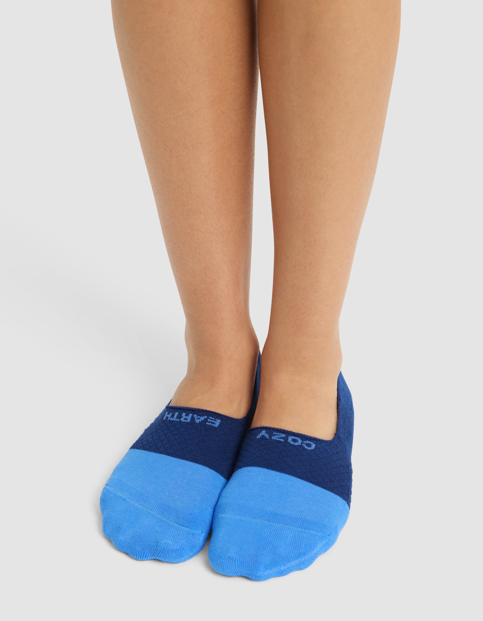 A person stands with feet together, wearing Cozy Earth Essential No-Show Socks in bright blue and navy with "COZY EARTH" displayed on top, against a plain white background. 