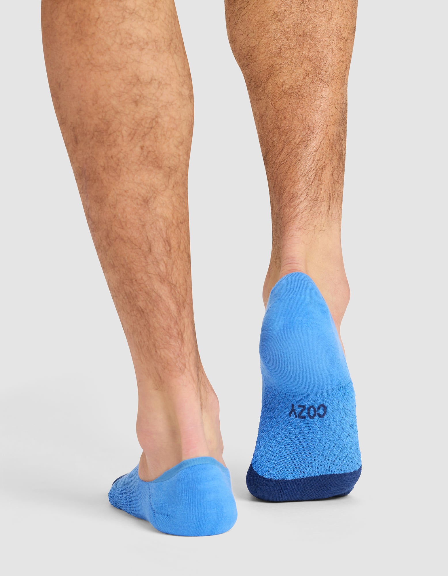 A person stands on a white background, viewed from behind, wearing bright blue Essential No-Show socks from Cozy Earth with "COZY" printed on the soles. 
