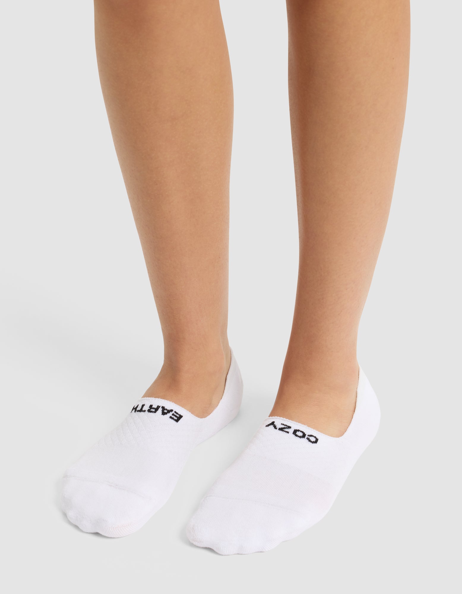 A person wearing Cozy Earth Essential No-Show Socks with "COZY" and "EARTH" in black on top stands on a light-colored surface. 