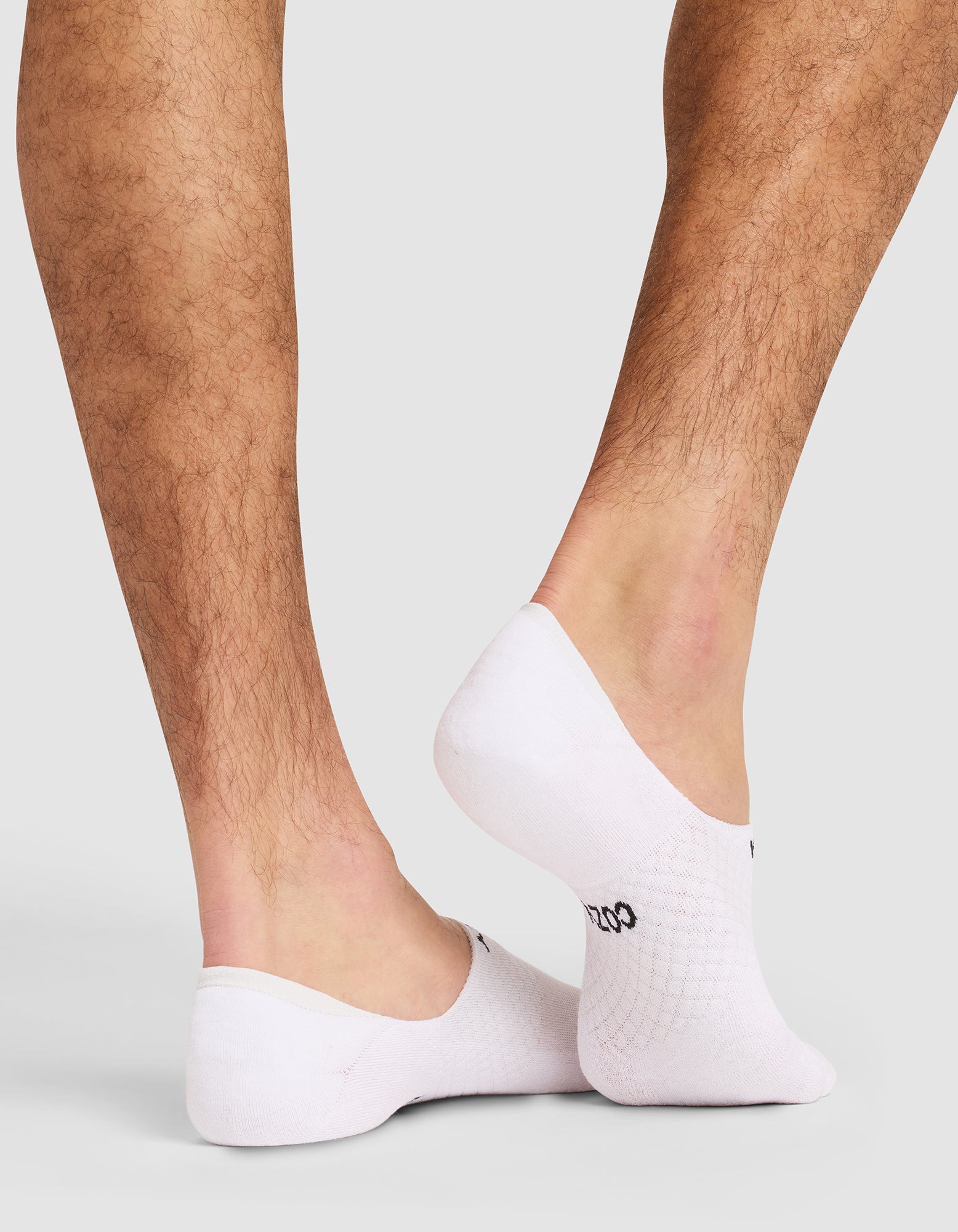 A close-up of a person's lower legs and feet showcases Cozy Earth's Essential No-Show Sock in white. The socks cover only the feet, leaving ankles and most of the legs bare against a light gray background. 