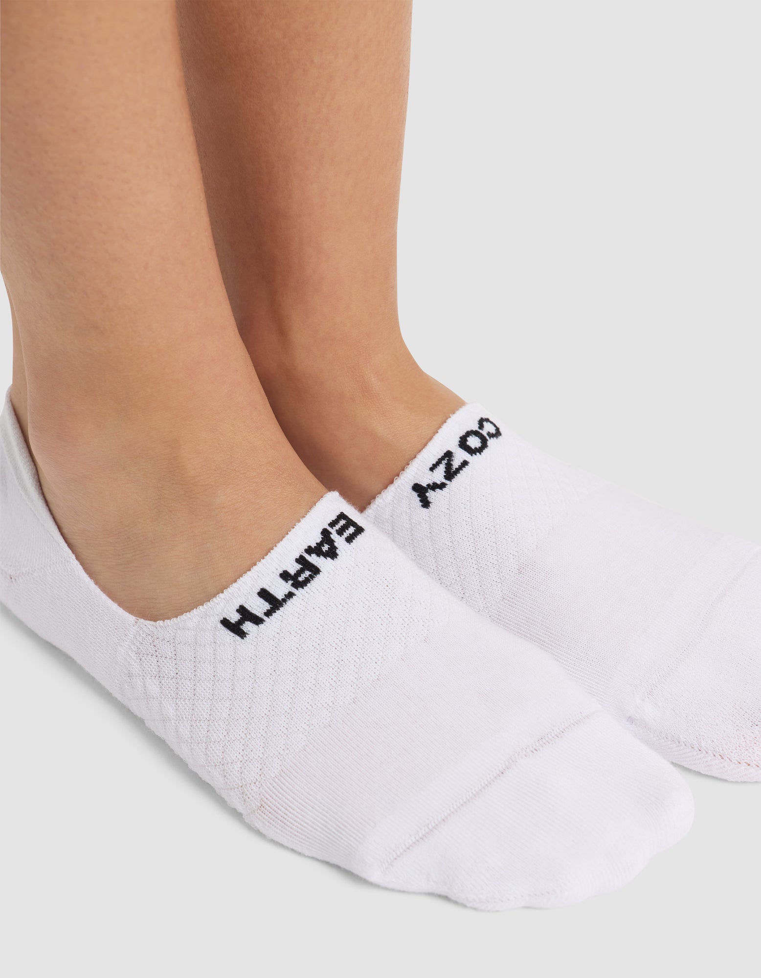 A close-up of two feet in Cozy Earth Essential No-Show Socks with black text—“EMPATH” on the left, “COZY” on the right—standing on a white surface. |Color:White