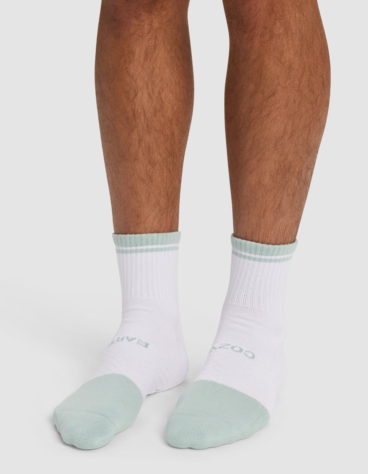 A person wearing Cozy Earth's Essential Quarter Sock - Last Chance stands on a white surface, showing only their lower legs and feet. The socks are white with light green accents on the toes, heels, and cuffs.